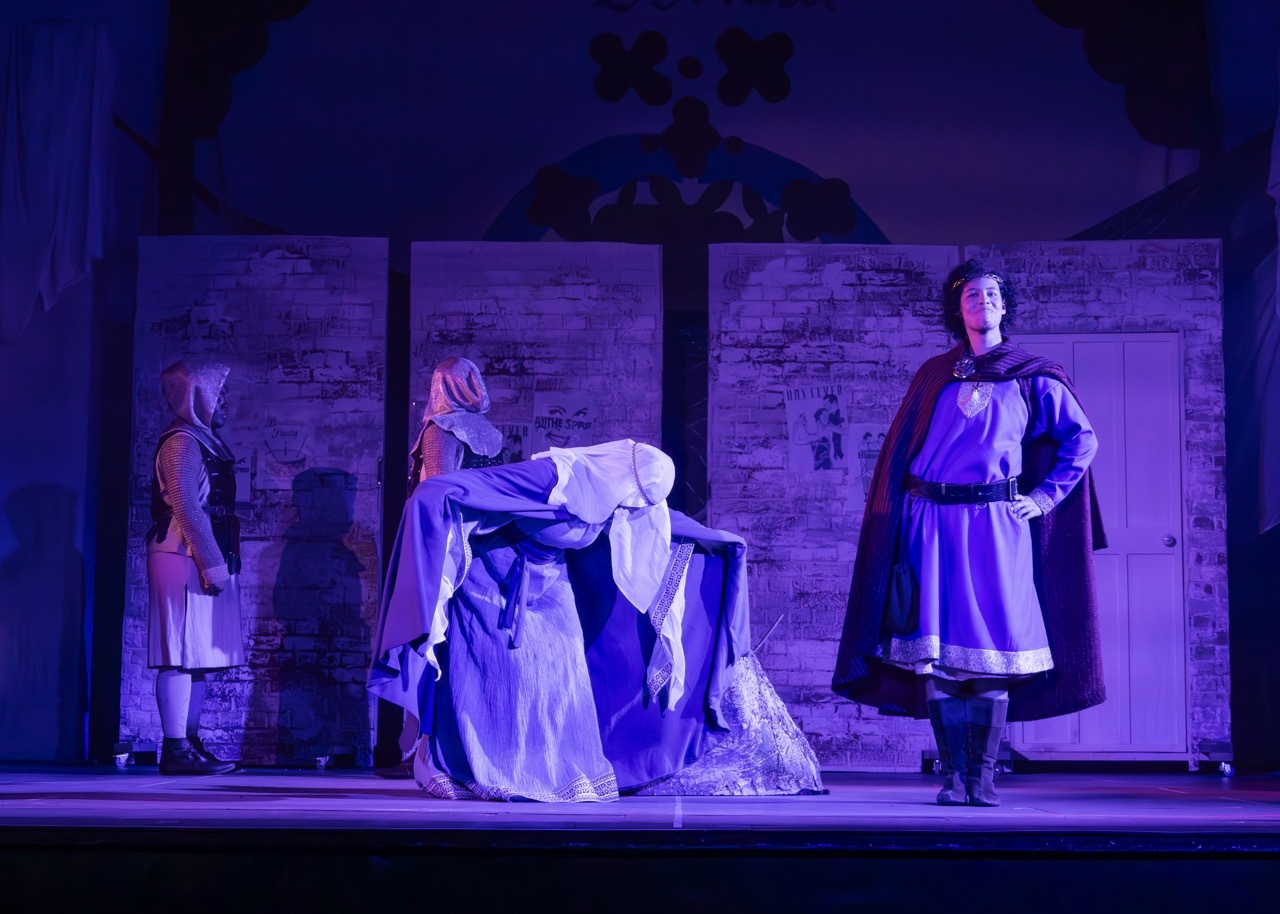 Theatre stage lit in purple and blue with four costumed performers in medieval robes against stone wall backdrop.