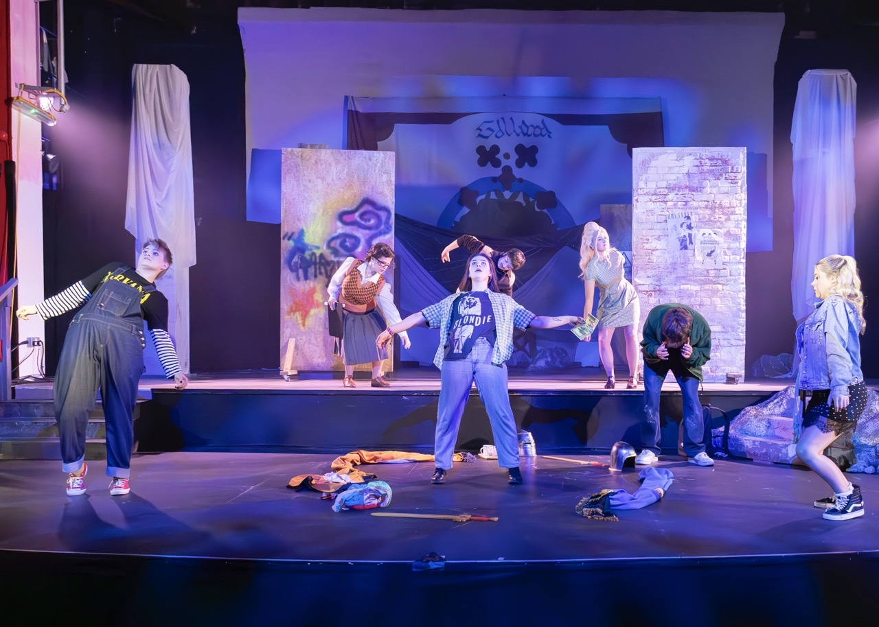 Theatre stage with actors in casual clothing performing. Blue and purple lighting illuminates backdrop with graffiti-style panels and white draped columns.
