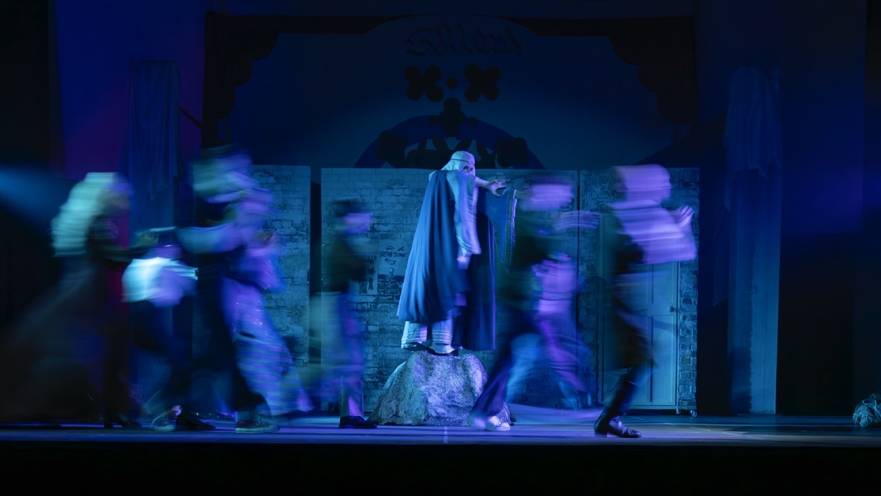 Theatre stage with blue lighting. Central figure in light blue robes stands on elevated rock. Blurred figures in motion on either side.
