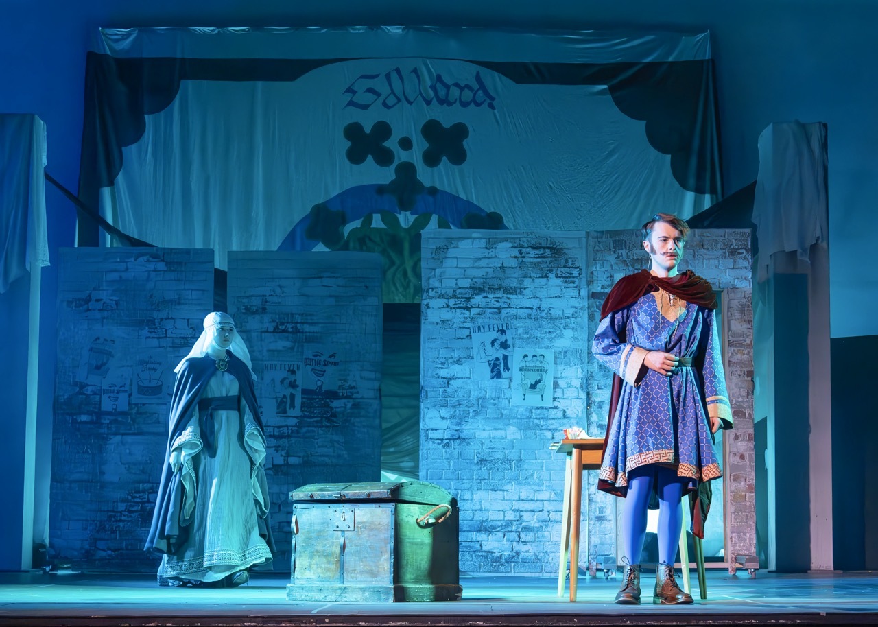 Theatre stage with blue lighting, two performers in medieval costumes, stone archway backdrop with decorative banners and wooden props.