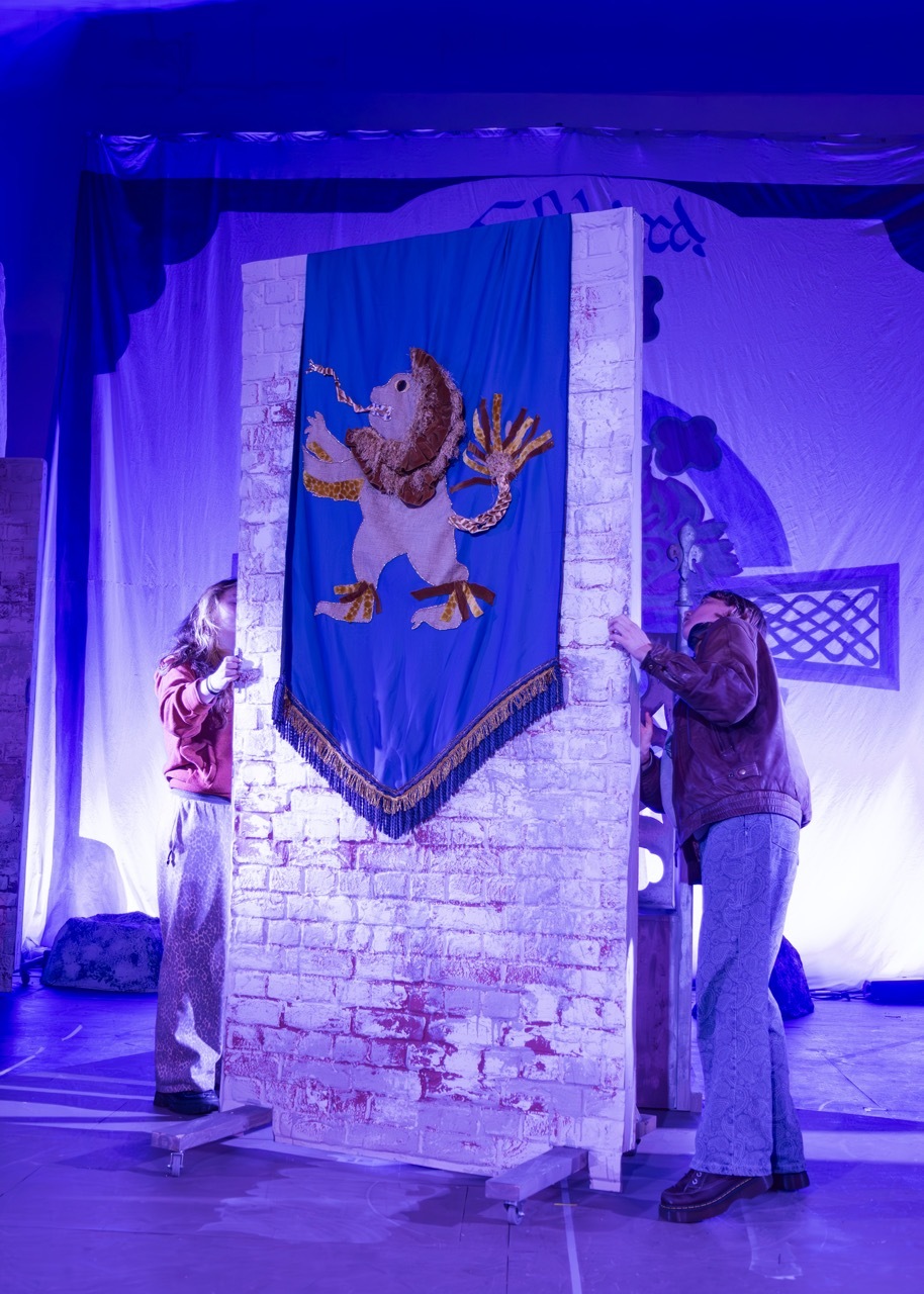 Theatre stage with blue and purple lighting. Two set designers beside a white brick tower with blue banner featuring brown character design.