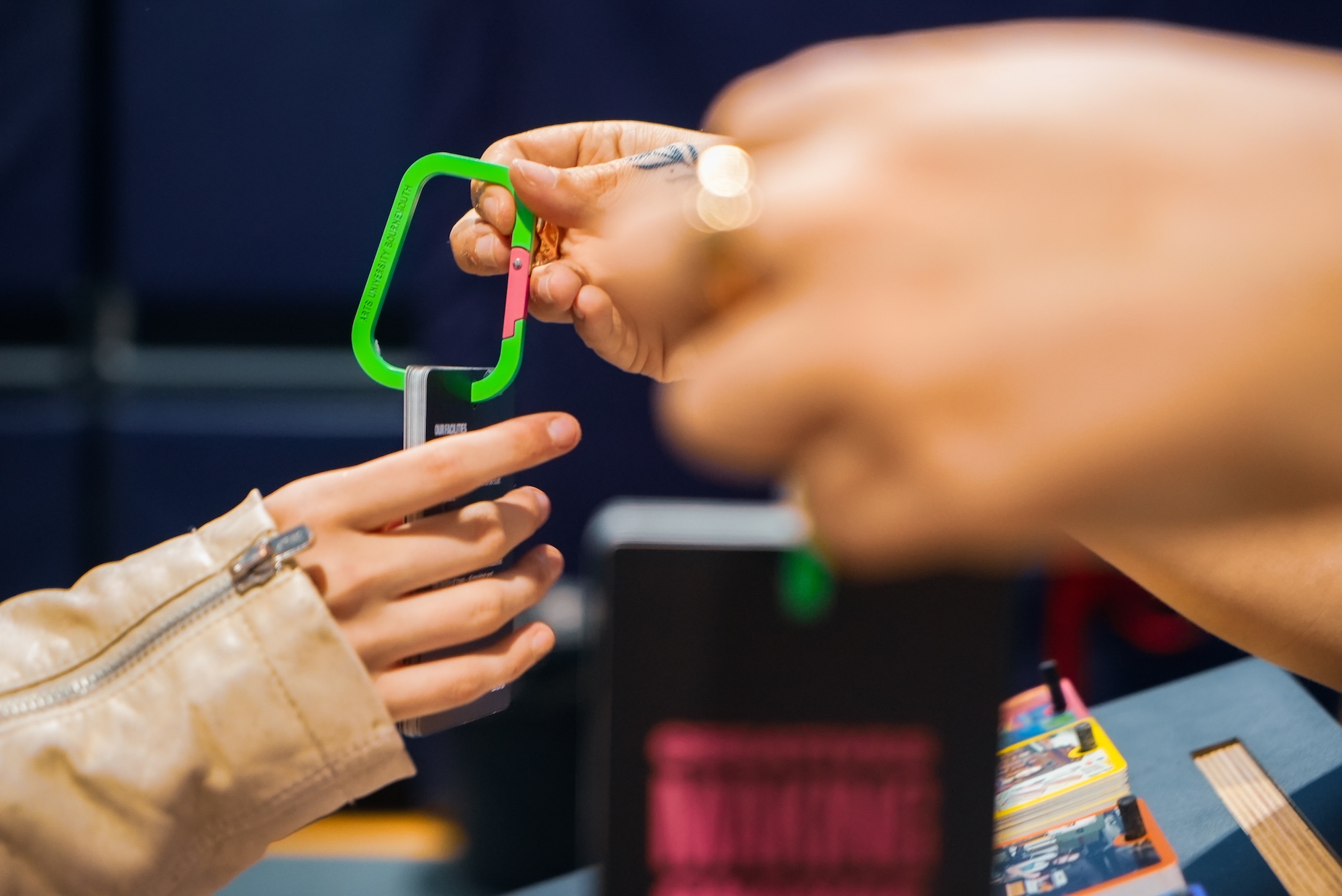 Hands holding and reaching out for a green-pink carabiner, blurred hands in front.