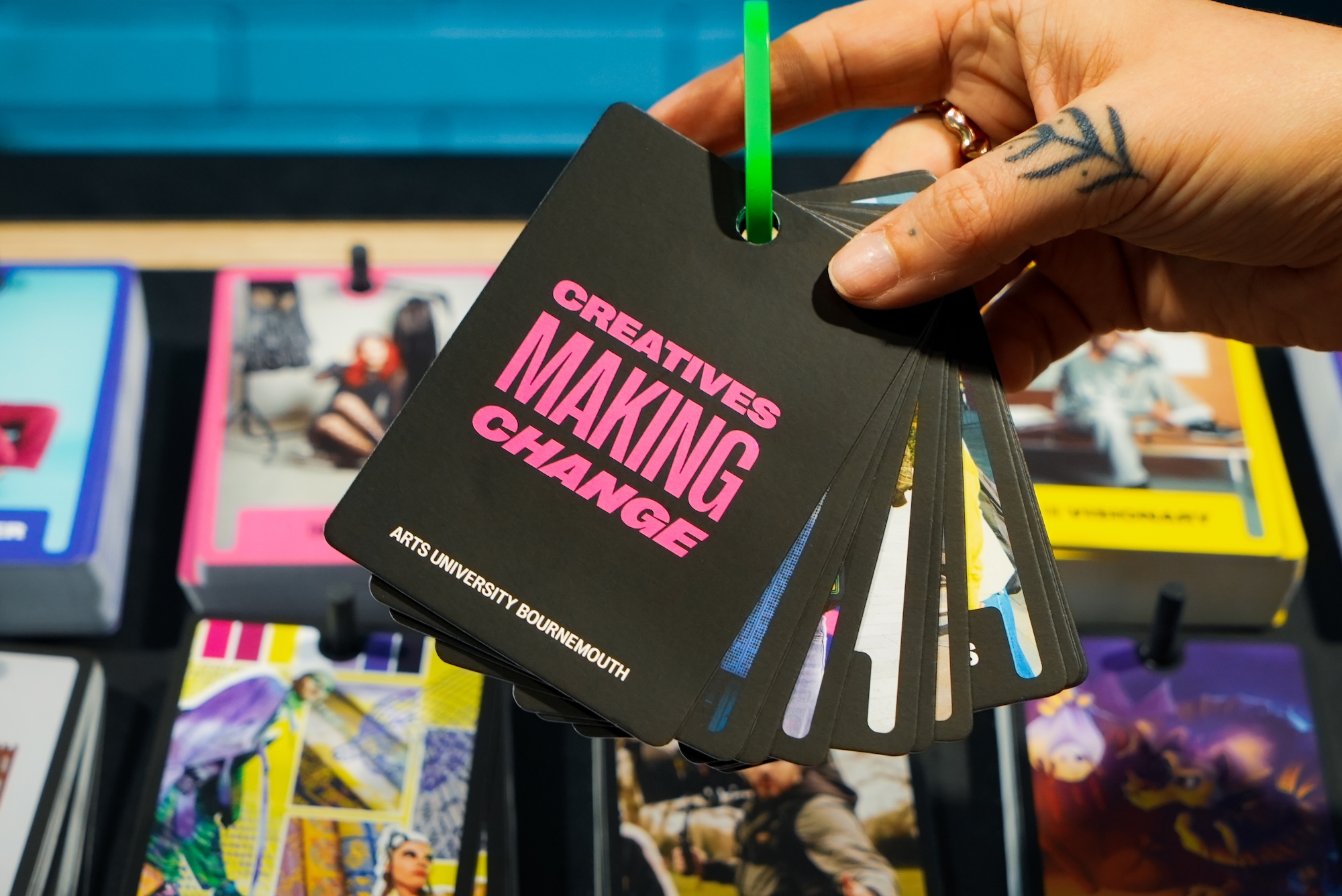 Black card with 'creatives making change' in pink text held by someone's hand, attached to a green-pink carabiner, other colourful cards in the background.