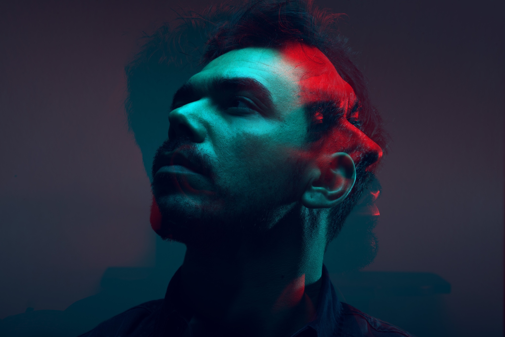 A close-up portrait of a man with dark hair, set against a gradient background. The image features a layered effect with shades of blue and red, creating a blurred and stylised visual appearance.