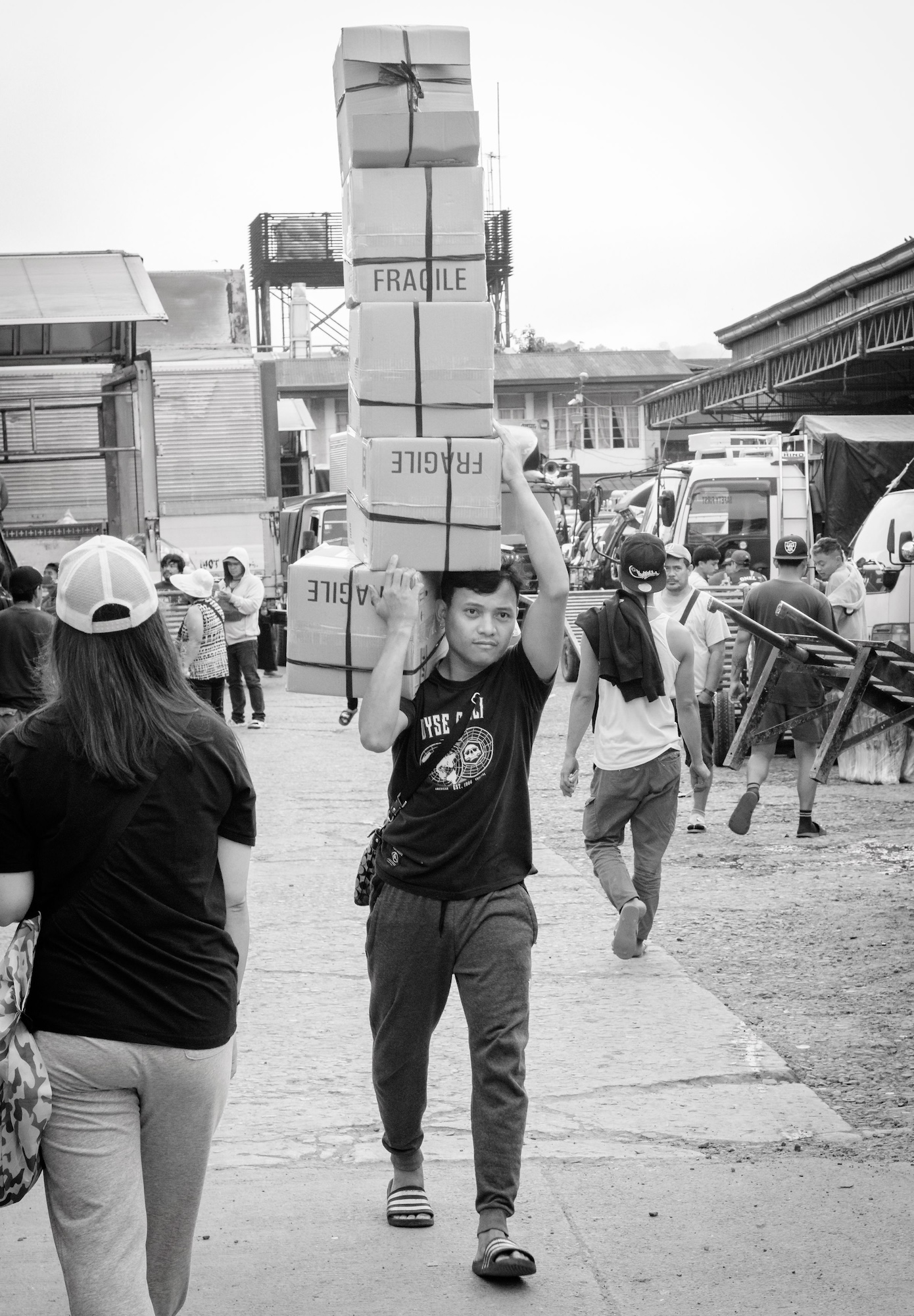 A young man in a black shirt and shorts carries a tall stack of cardboard boxes marked 'FRAGILE' on his head. People walk in the background along a concrete pathway beside vehicles and industrial buildings.