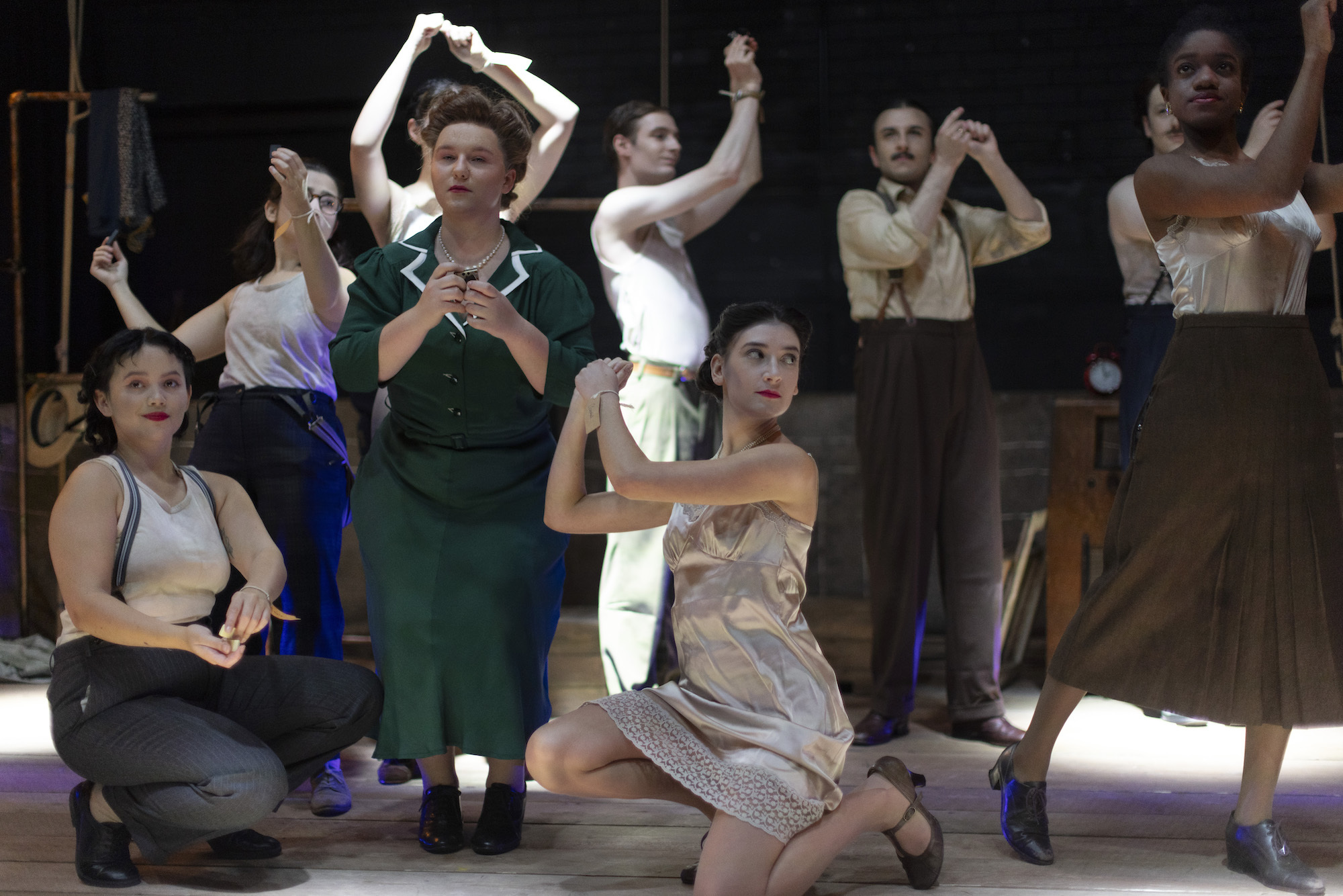 A group of BA (Hons) Acting students in 1940s style costume on stage performing "Thérèse". Most performers are standing with their arms raised. Two performers are crouched down in the foreground.