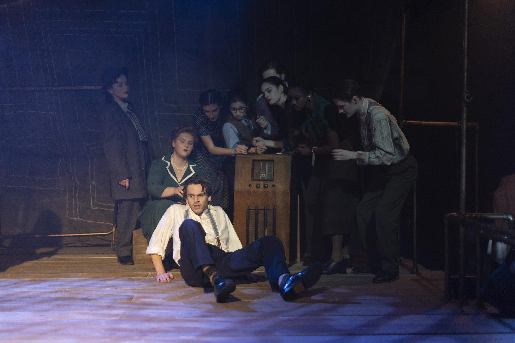 A group of BA (Hons) Acting students on stage performing "Thérèse". One performer is on their backside, cowering back towards an old-fashioned radio, while the other performers are gathered around it.
