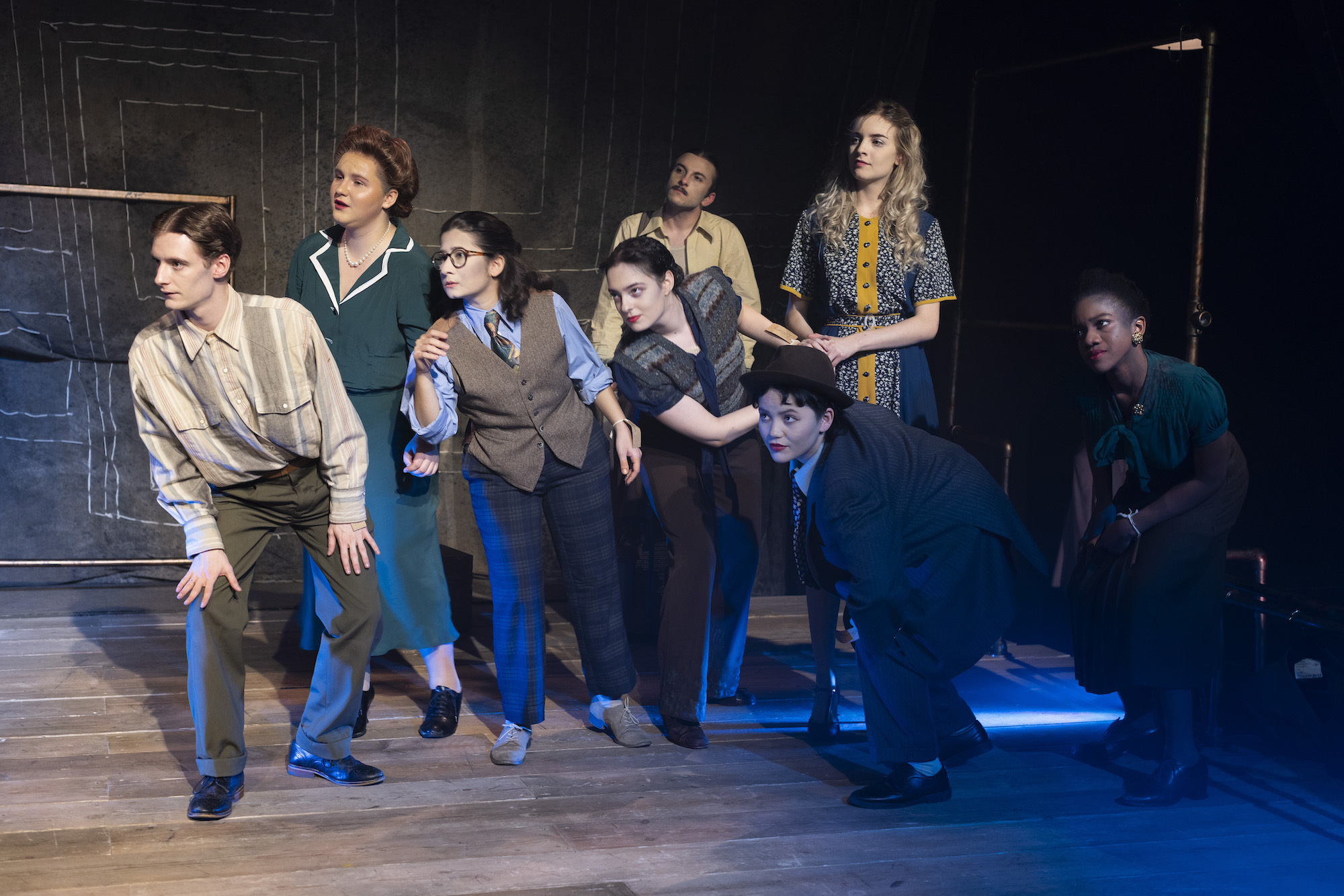 A group of BA (Hons) Acting students dressed in 1940s style costume on stage performing "Thérèse". All performers are looking to the left and leaning in as if listening.