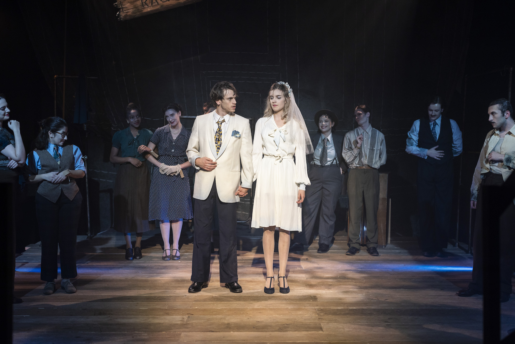 Two BA (Hons) Acting students standing in the centre of a stage under a spotlight, holding hands. One is wearing a bridal veil. Other performers are standing in partial darkness around the edges of the stage.