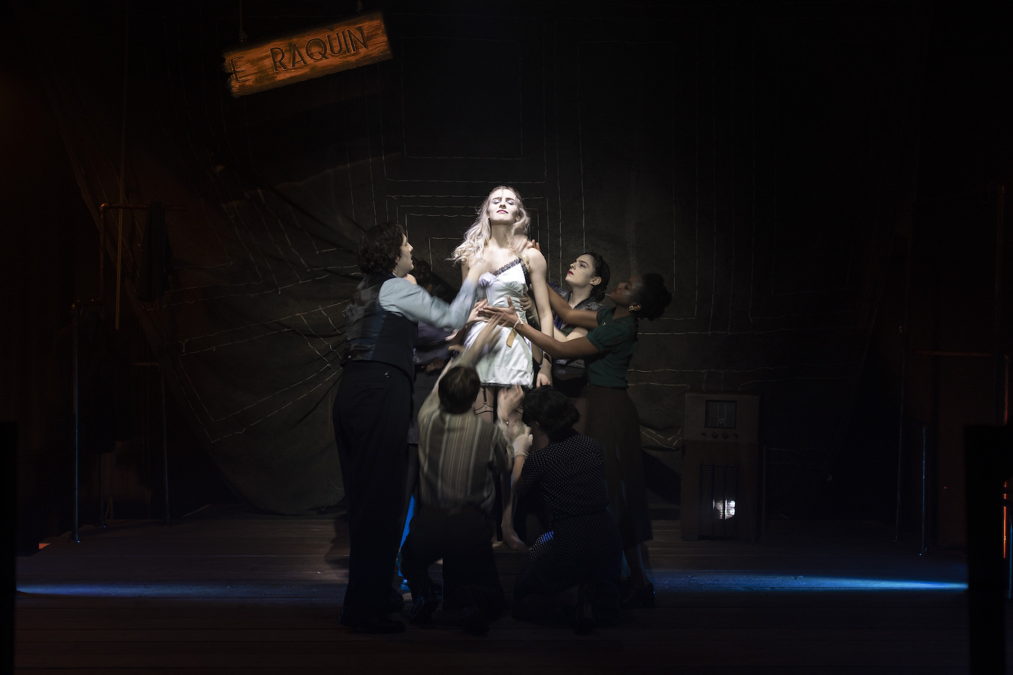 A BA (Hons) Acting student standing on stage performing "Thérèse" wearing a negligee under a spotlight. A small group of fellow performers are gathered around her, holding her by the waist and shoulders.