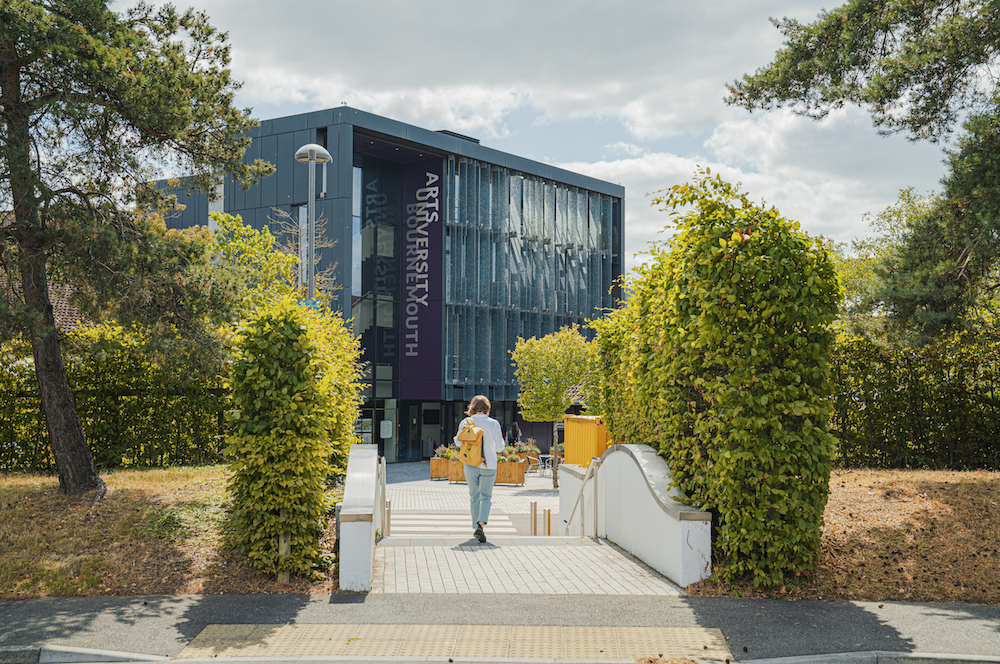 Modern dark grey building with vertical text, person walking up white steps flanked by green hedges and trees under cloudy sky.