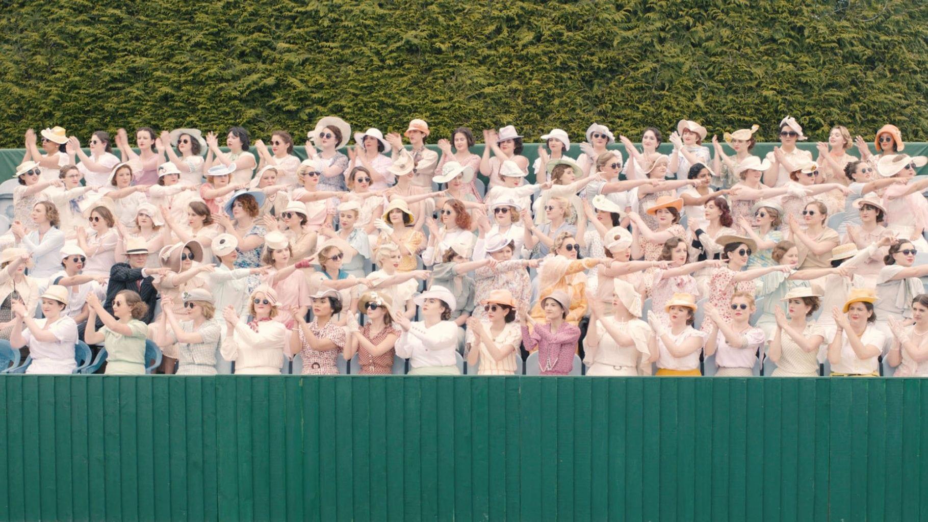 A large audience in period costume sitting in stands watching a tennis match.