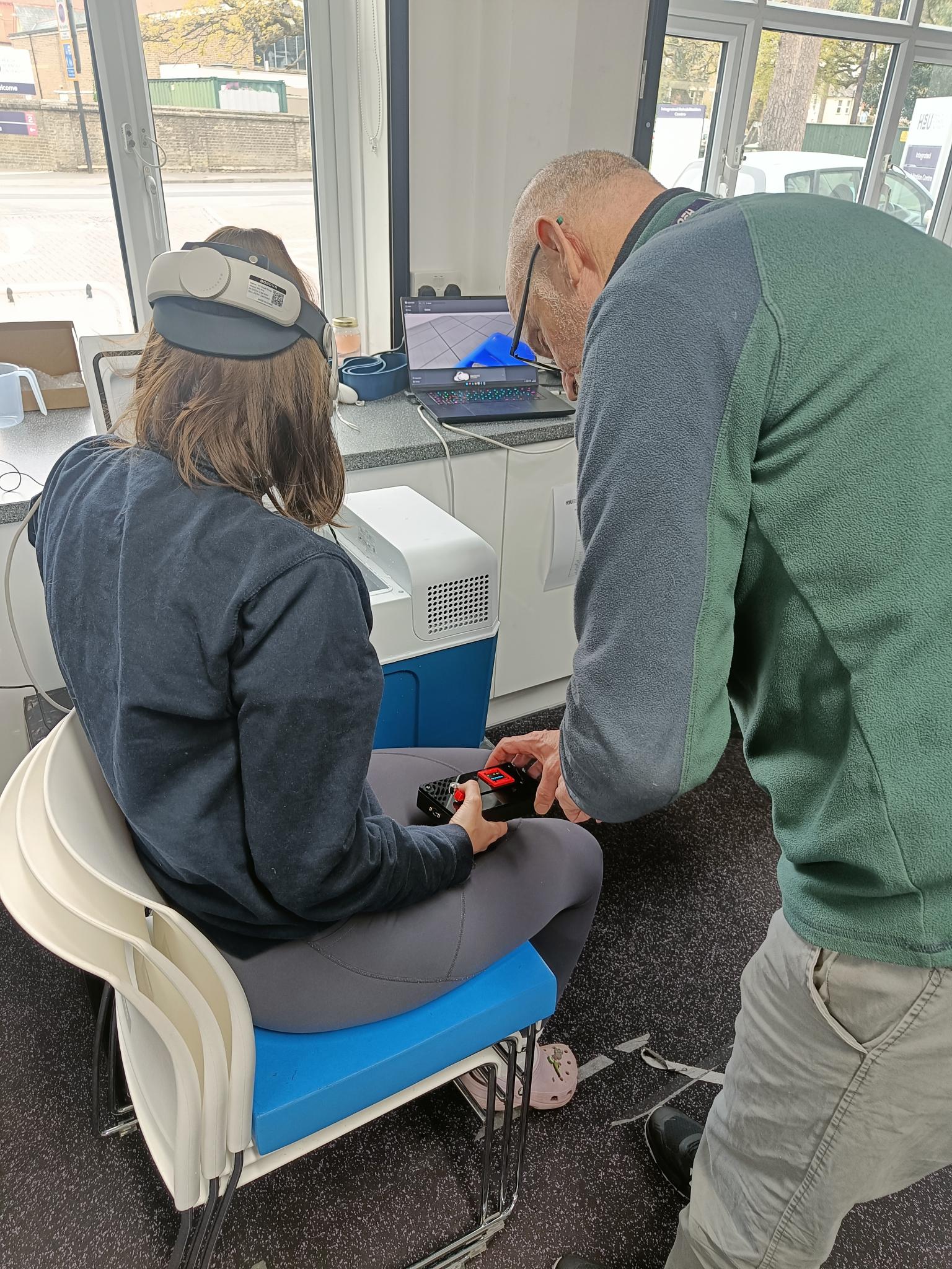 A person wearing a grey sweater and a headset sits on a chair, while a person with glasses and a green sweater adjusts equipment on her leg. In the background, there are tables with a computer and a window showing outdoor views.