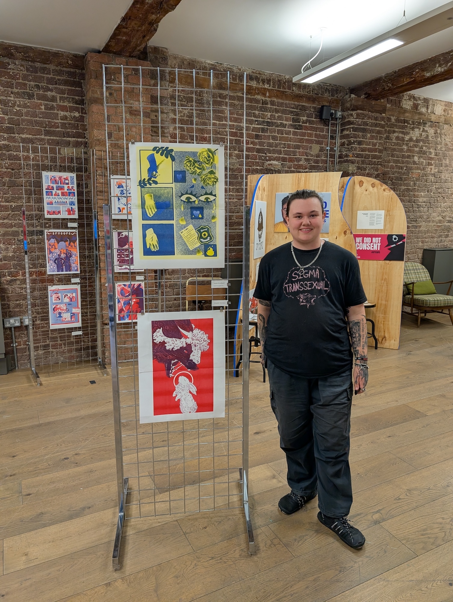 Man in black t-shirt standing beside metal display rack with colourful artwork in brick-walled gallery space with exposed beams.