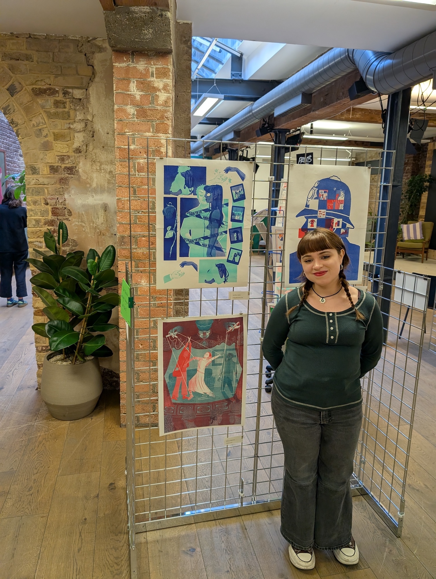 Woman in green top and trousers standing beside metal display grid with colourful artwork in brick-walled gallery space.