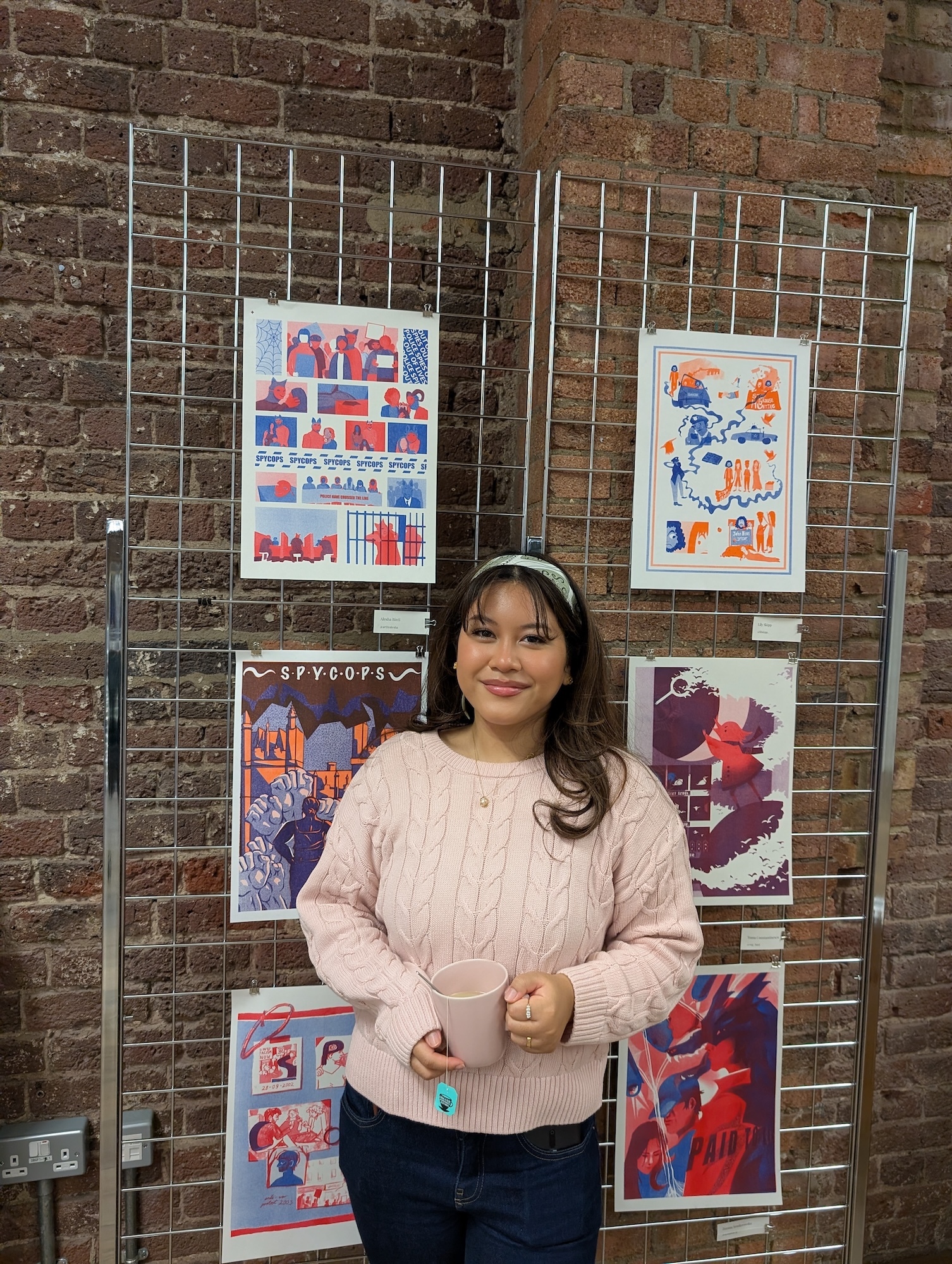 Woman in beige jumper stands before metal display rack with colourful artwork prints against brick wall.