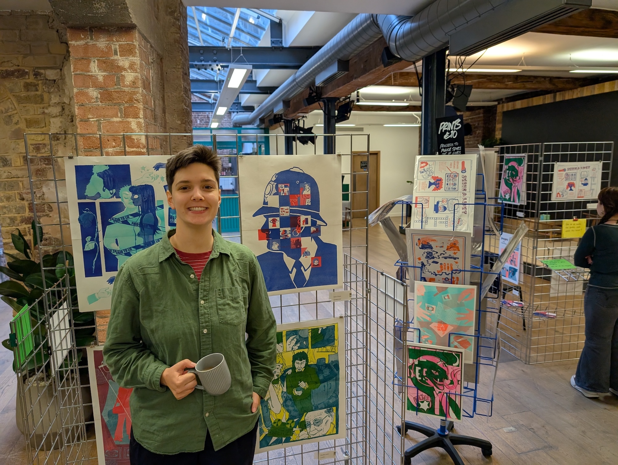 Woman in green shirt standing beside wire display racks filled with colourful artwork in industrial-style space with exposed brick walls.