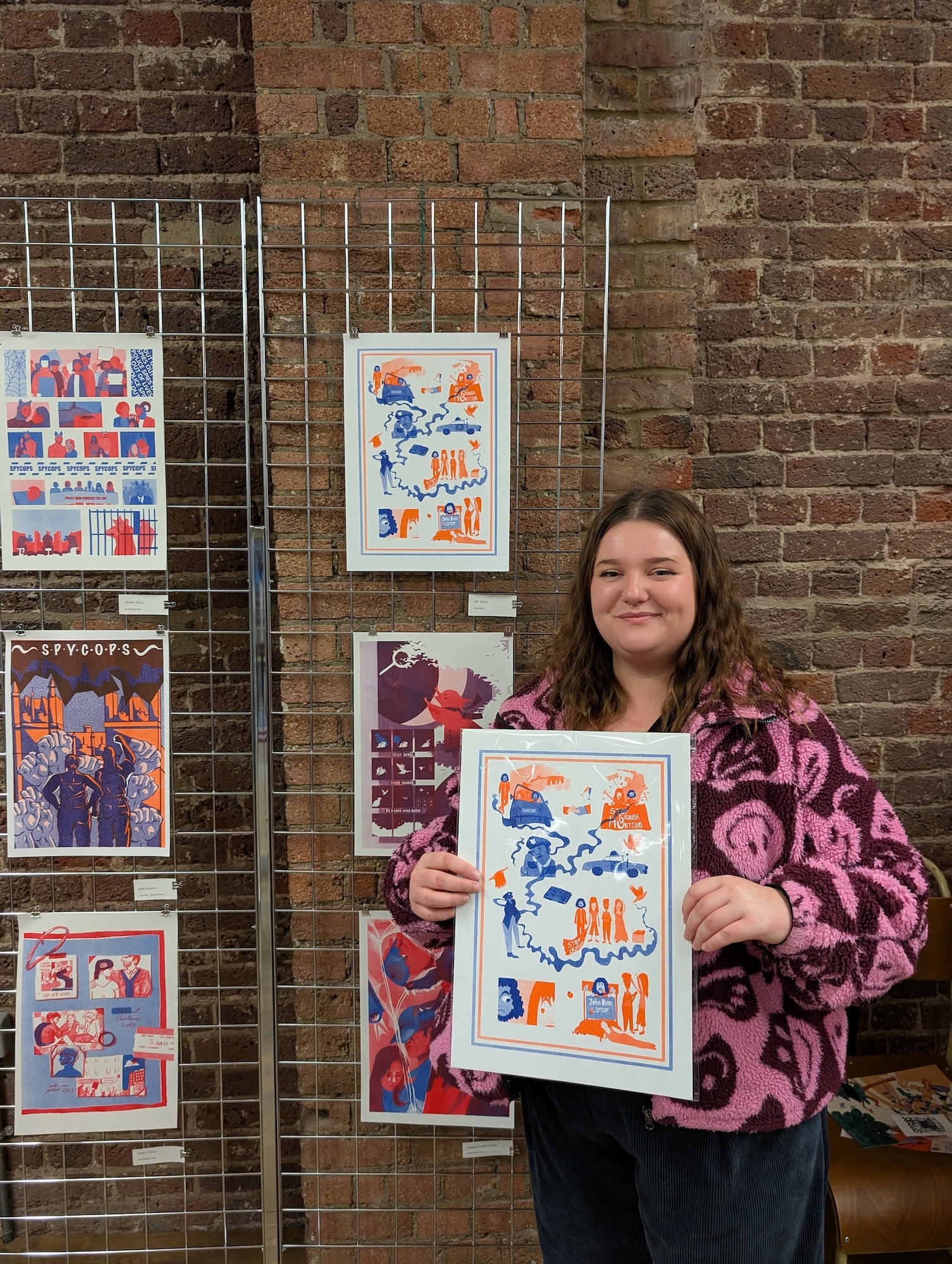 Woman in pink patterned top holding framed artwork displaying blue and orange illustrations, standing beside wire display rack with various colourful prints against brick wall.