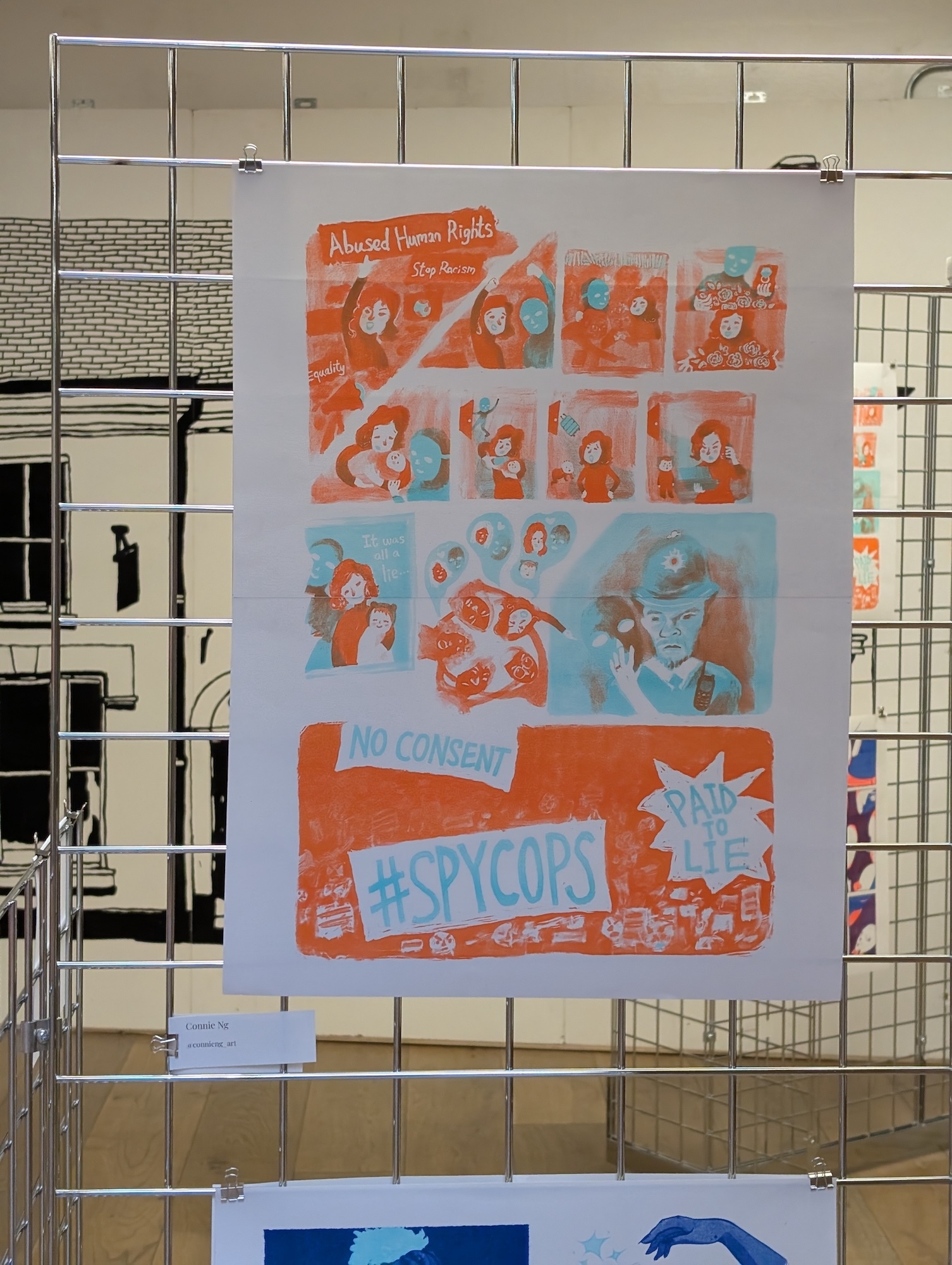 Colourful children's artwork displayed on metal grid. Orange and blue poster with cartoon characters, text reads "SPYCOPS" at bottom.