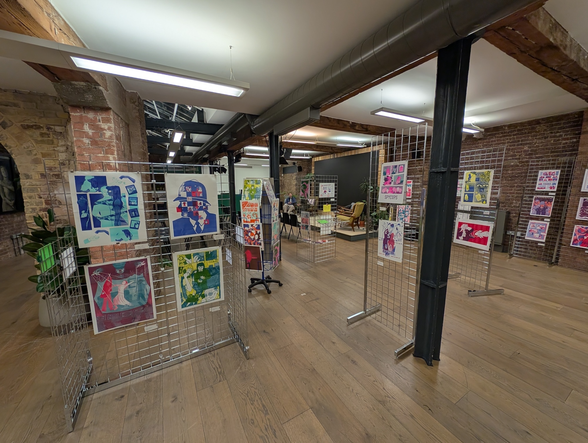 Industrial space with exposed brick walls and wooden floors displaying colourful artwork on metal wire display stands throughout the room.