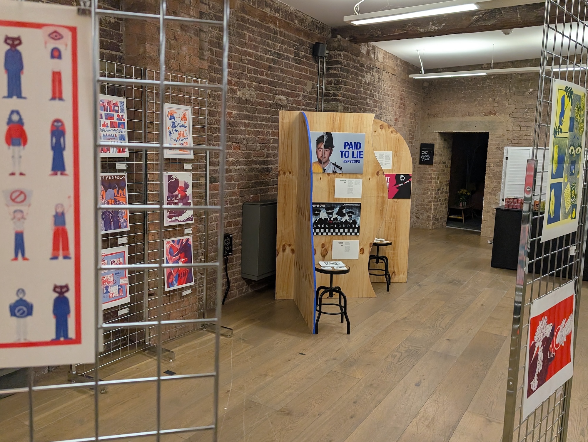 Exhibition space with wire display stands showing colourful posters, exposed brick walls, concrete floor, and wooden partition wall.
