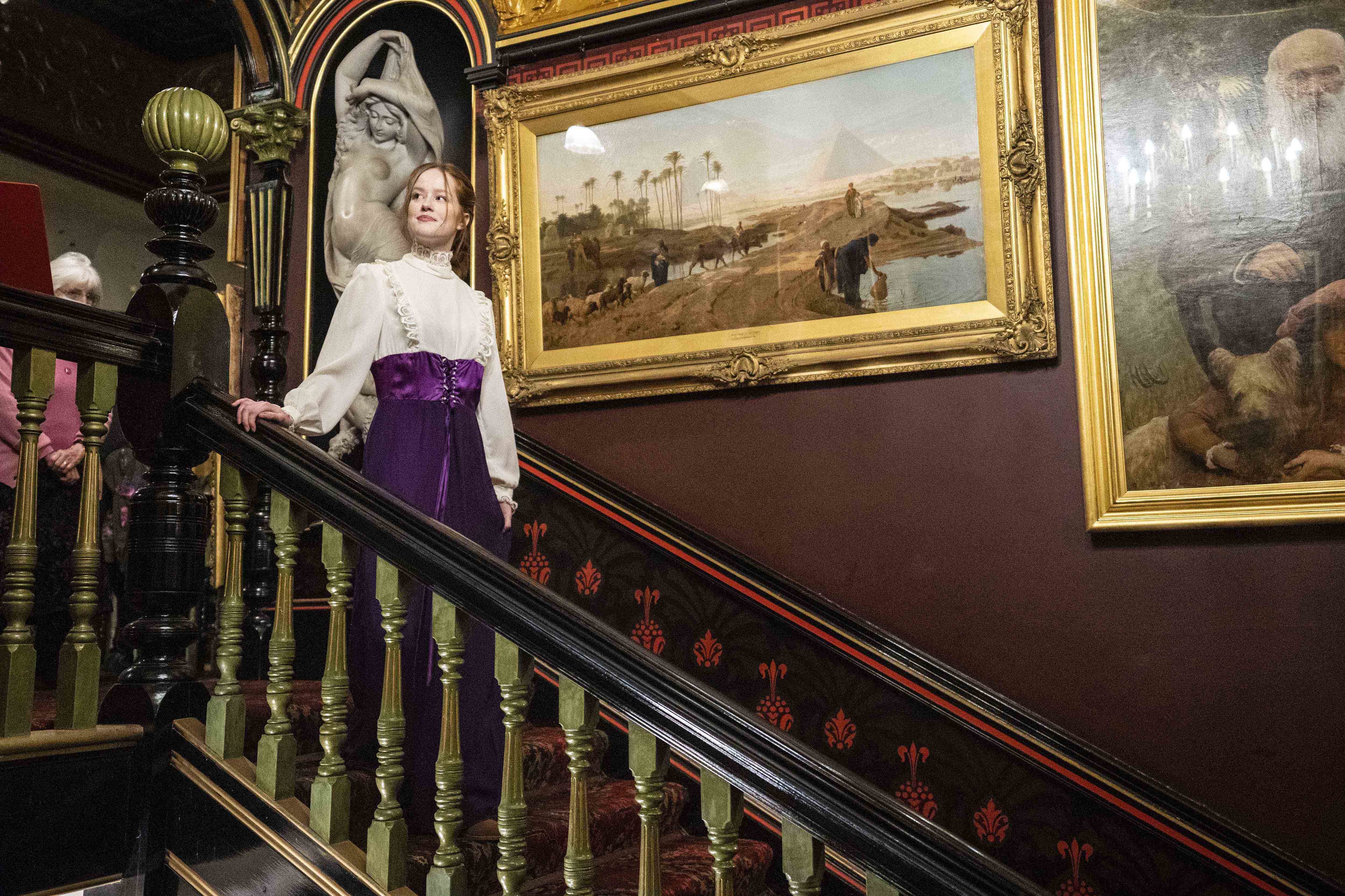 A person in period costume walks down a staircase in a mansion. There are gold-framed paintings on the walls.