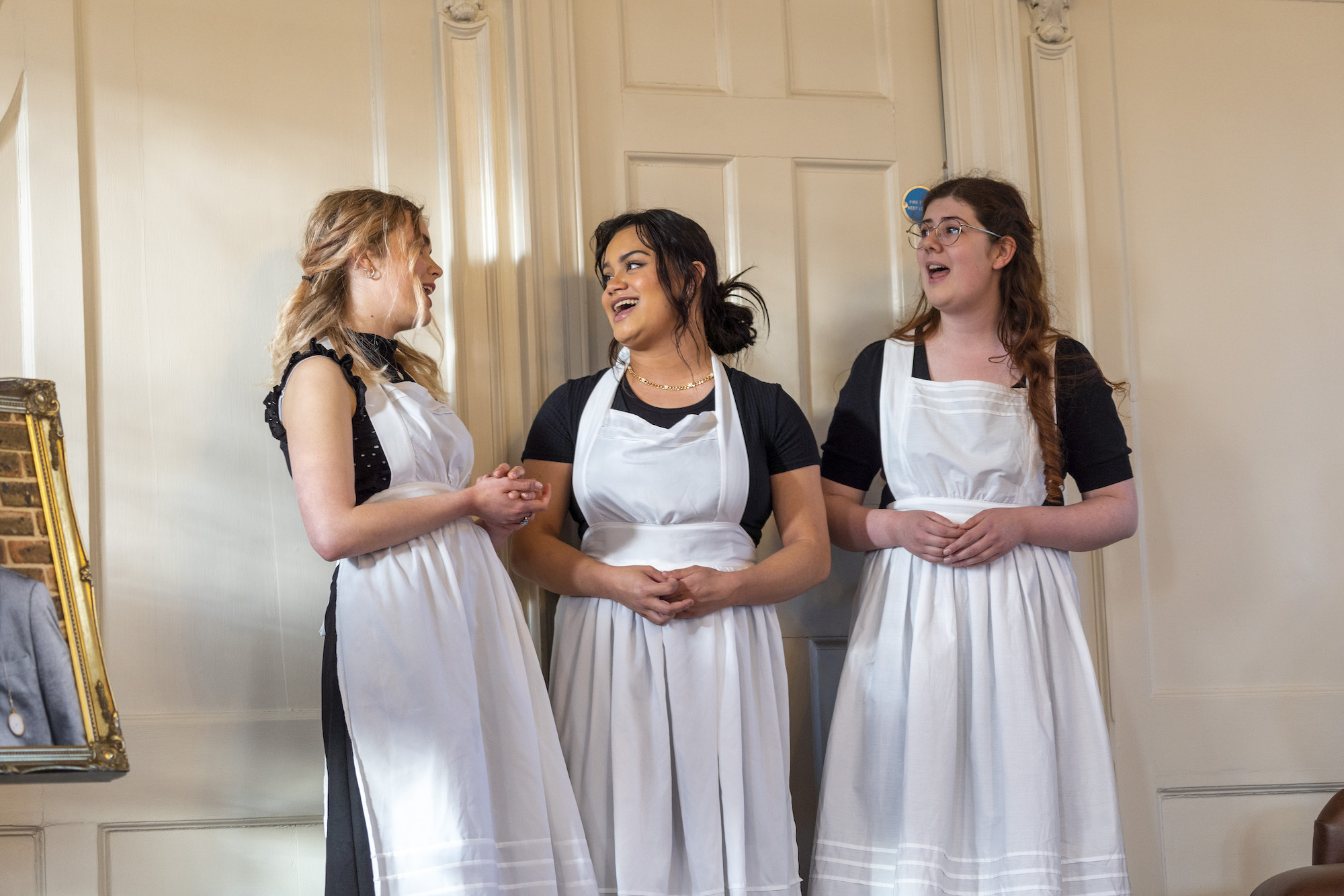 Three performers in long white aprons singing by a door.