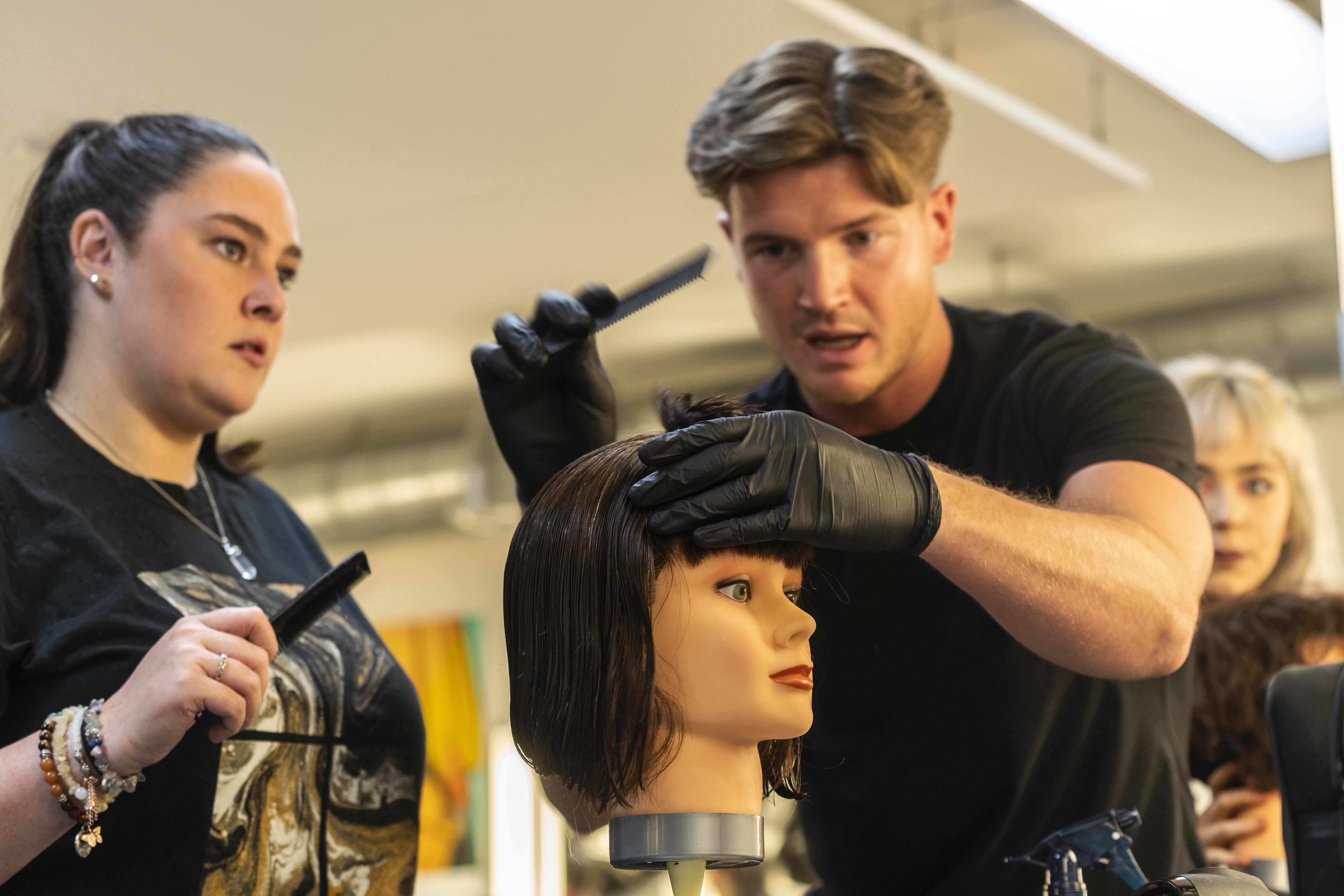 A barber holds a comb in the middle of styling the hair on a mannequin, while another person stands and observes.