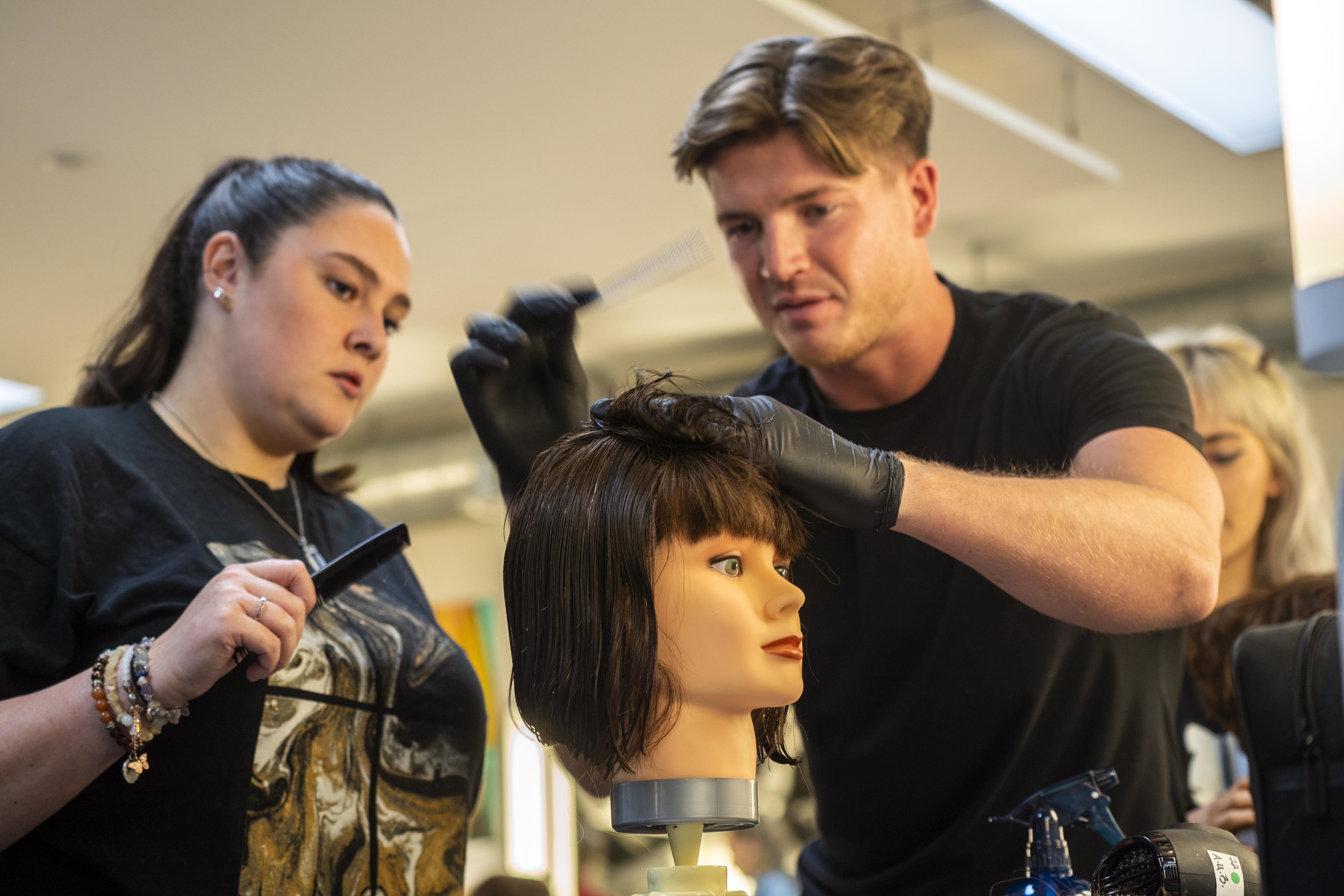 A barber holds a comb in the middle of styling the hair on a mannequin, while another person stands and observes.