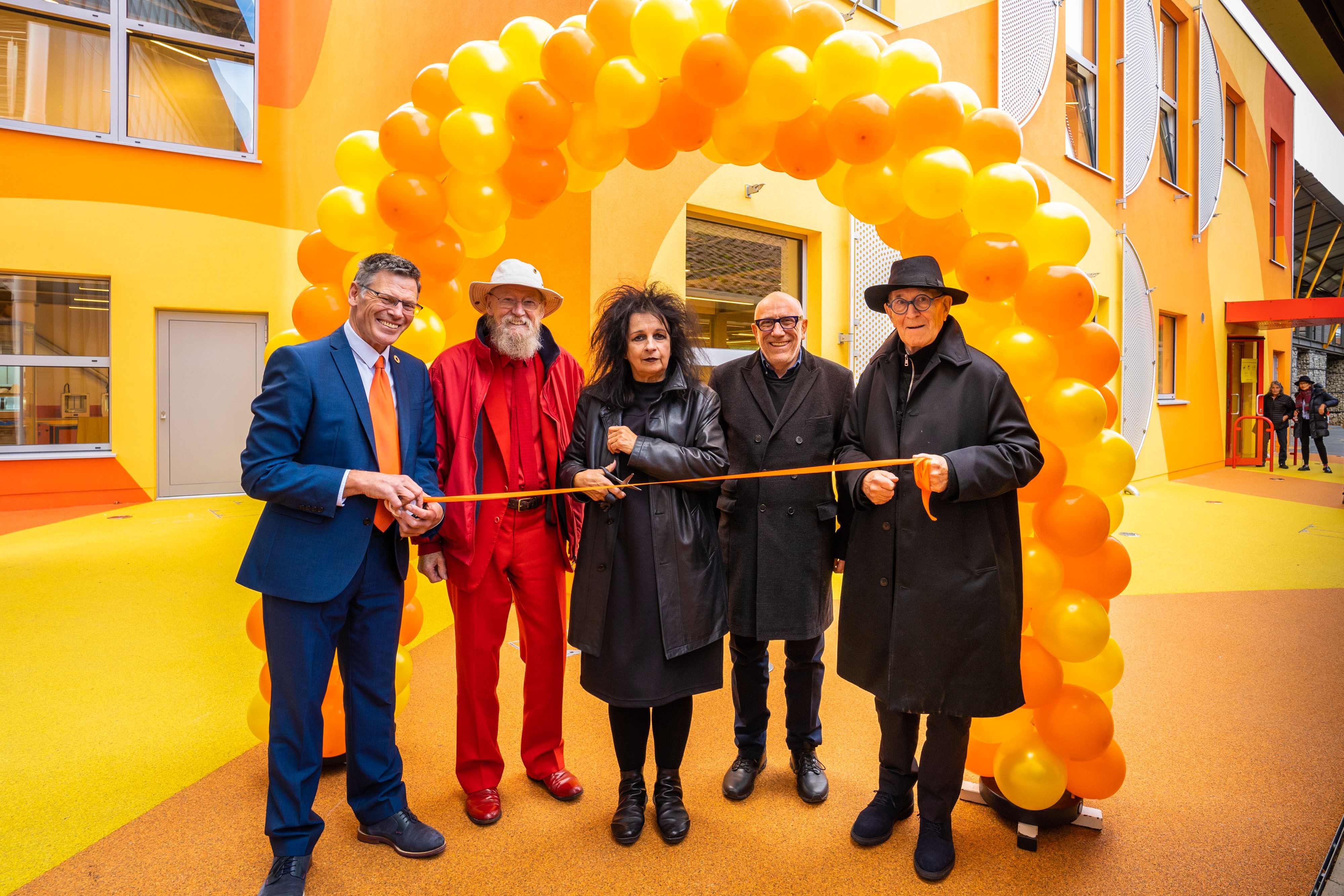 Five people stand outside a large orange building and under a balloon arch, holding a ribbon. Architect Odile Decq (Centre) opens AUB's new Innovation Studio with Vice-Chancellor Professor Paul Gough, Pompidou Centre Architect Mike Davies CBE, AUB Honorary Fellow Brian Clarke, and Innovation Studio Architect Professor Sir Peter Cook