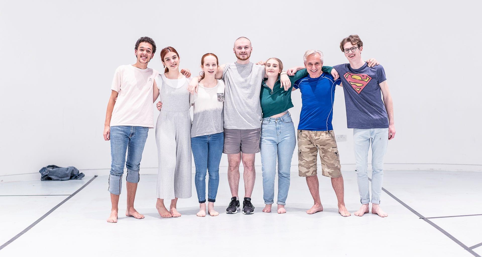 A group of seven people line up in a large white room and pose for a photo with their arms around each other.