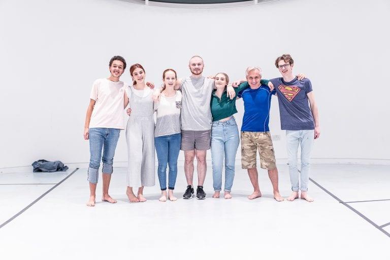 Smaller image of a group of seven people line up in a large white room and pose for a photo with their arms around each other.