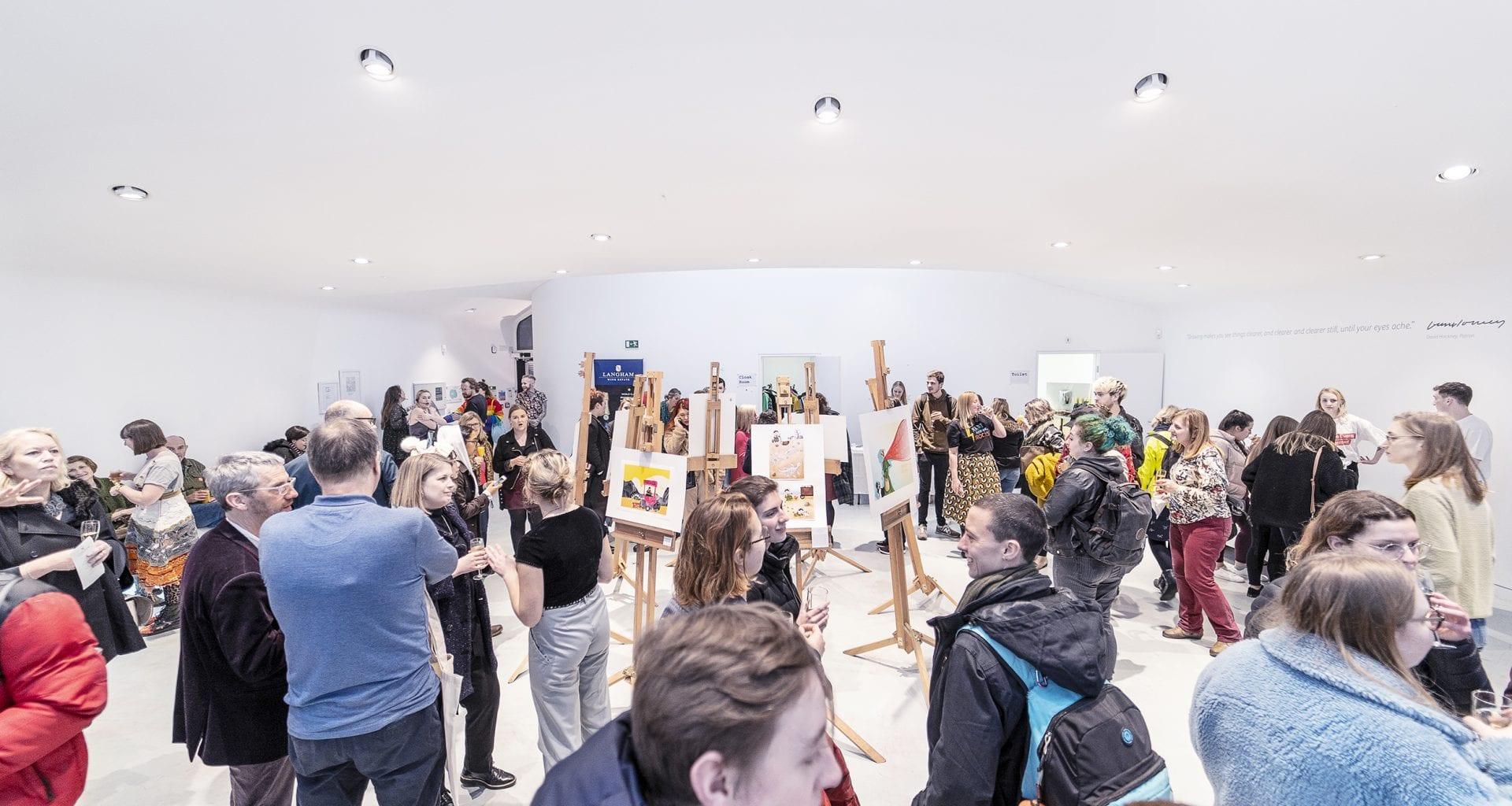 A crowd of people gathers in an indoor gallery space. Several pictures are on display stands in the middle of the room.