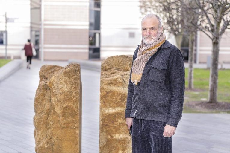 T Harrison standing in a courtyard with a pair of stone sculptures behind him.