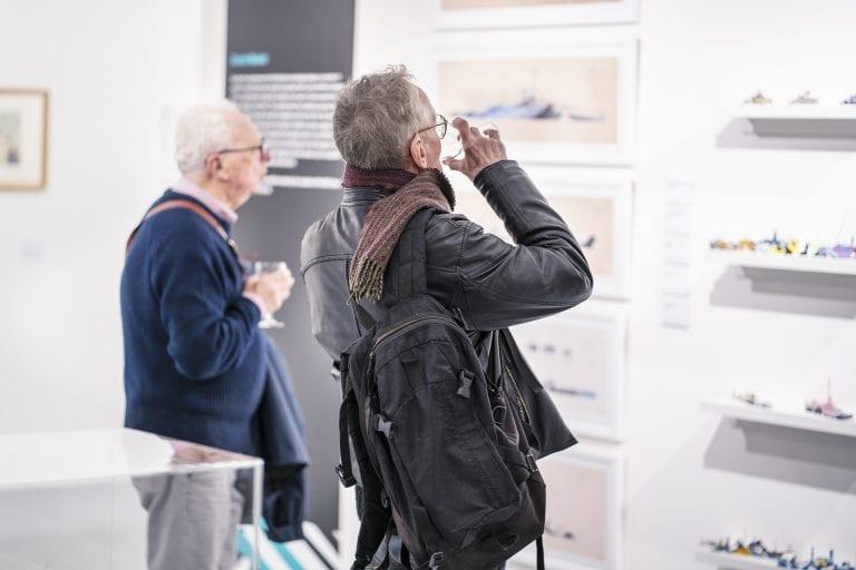 Two people holding drinks glasses look at a gallery exhibit at an event.