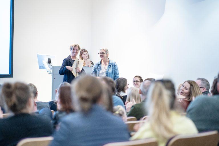 Three people stand at the front of a lecture theatre accepting an award in front of a large audience.