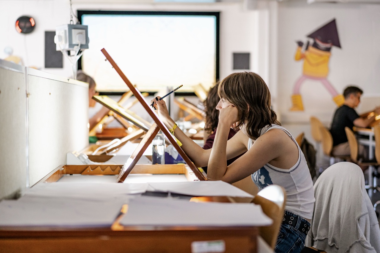 A young woman sketches at an easel in a bright art classroom, with tables, papers and other students in the background.