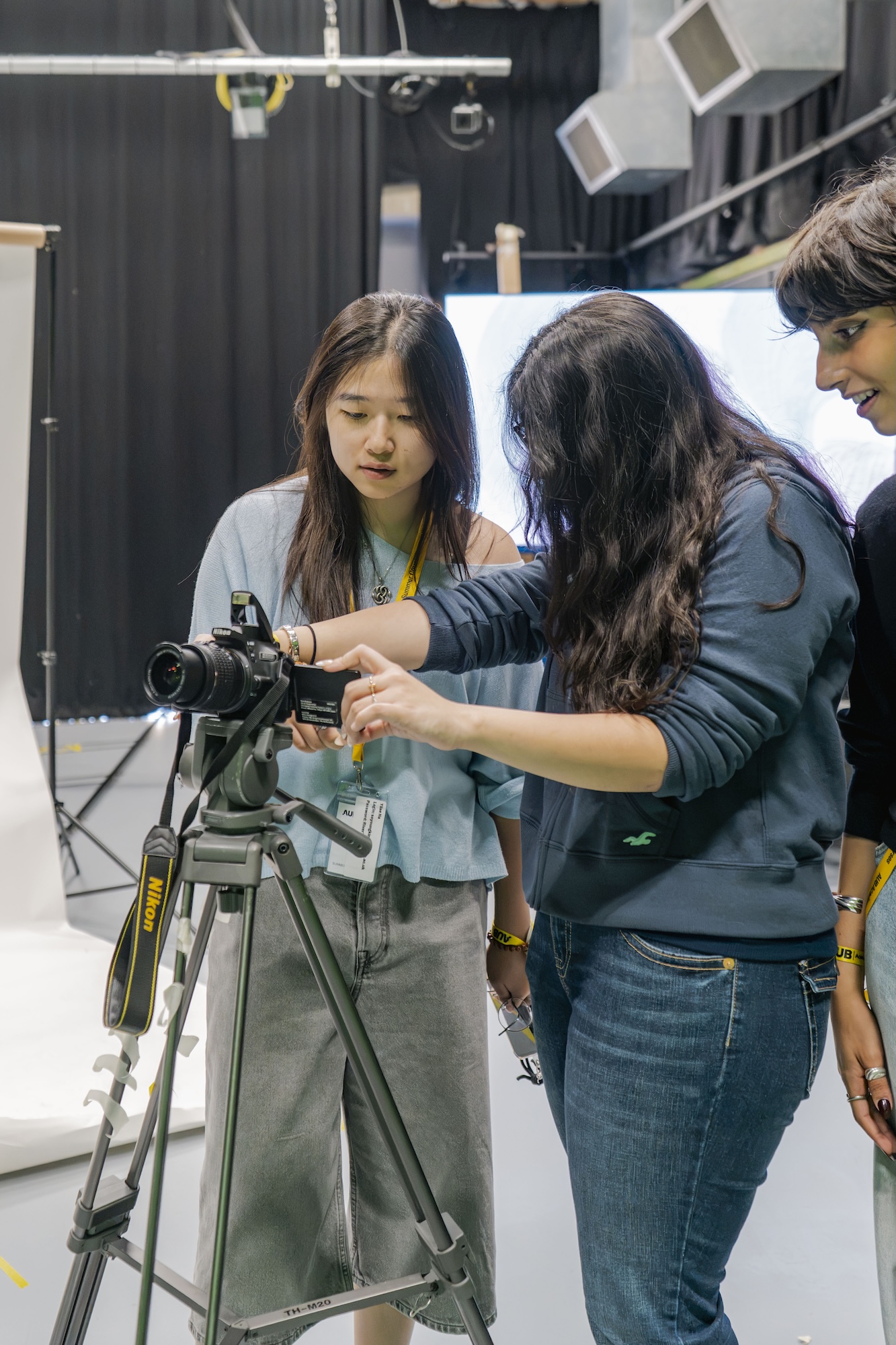 Three young women inspect a camera mounted on a tripod in a studio, with lighting equipment and black curtains in the background.