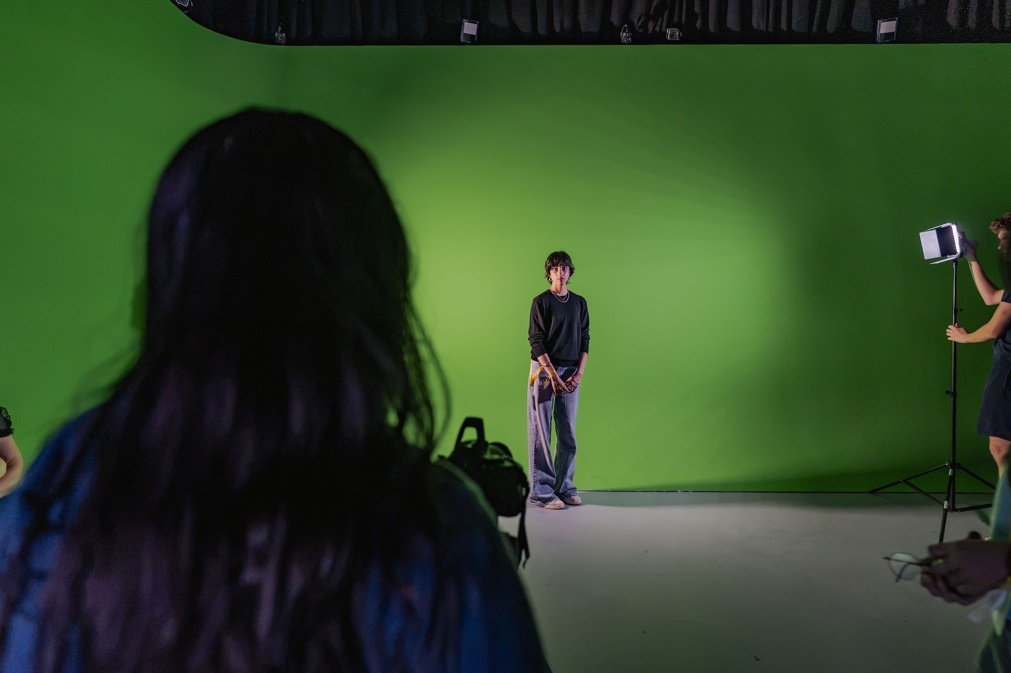 A young man stands in front of a green screen in a studio, with crew members and a lighting stand visible at the edges.
