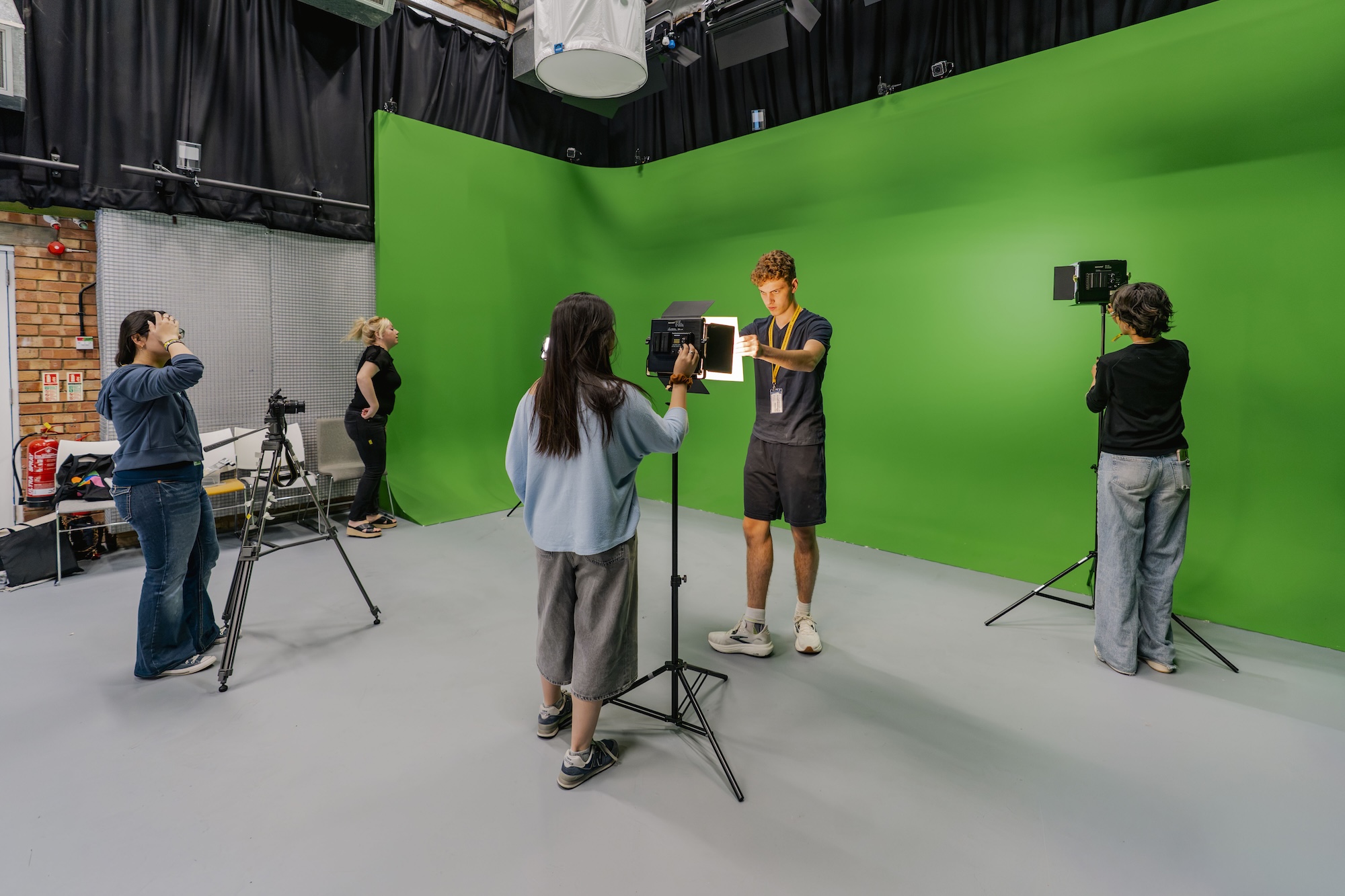Several people film a tall man in a studio with a green screen, using cameras and lighting stands on a grey floor.