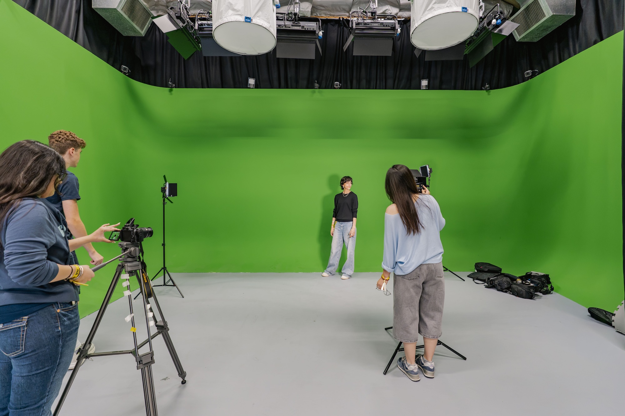 A film crew records a person standing before a large green screen in a studio, with cameras, lights and tripods visible around the set.