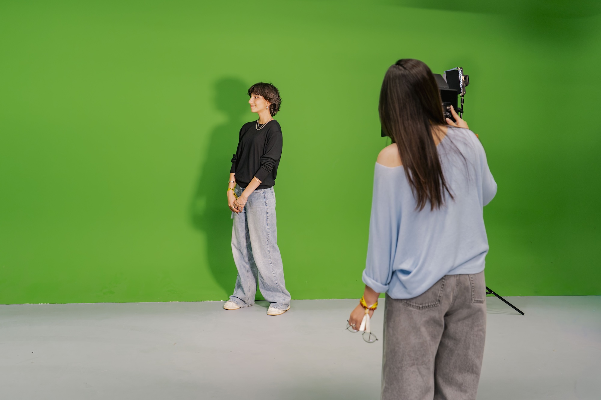A person photographs a model standing against a green studio backdrop, with a light grey floor and camera tripod visible.