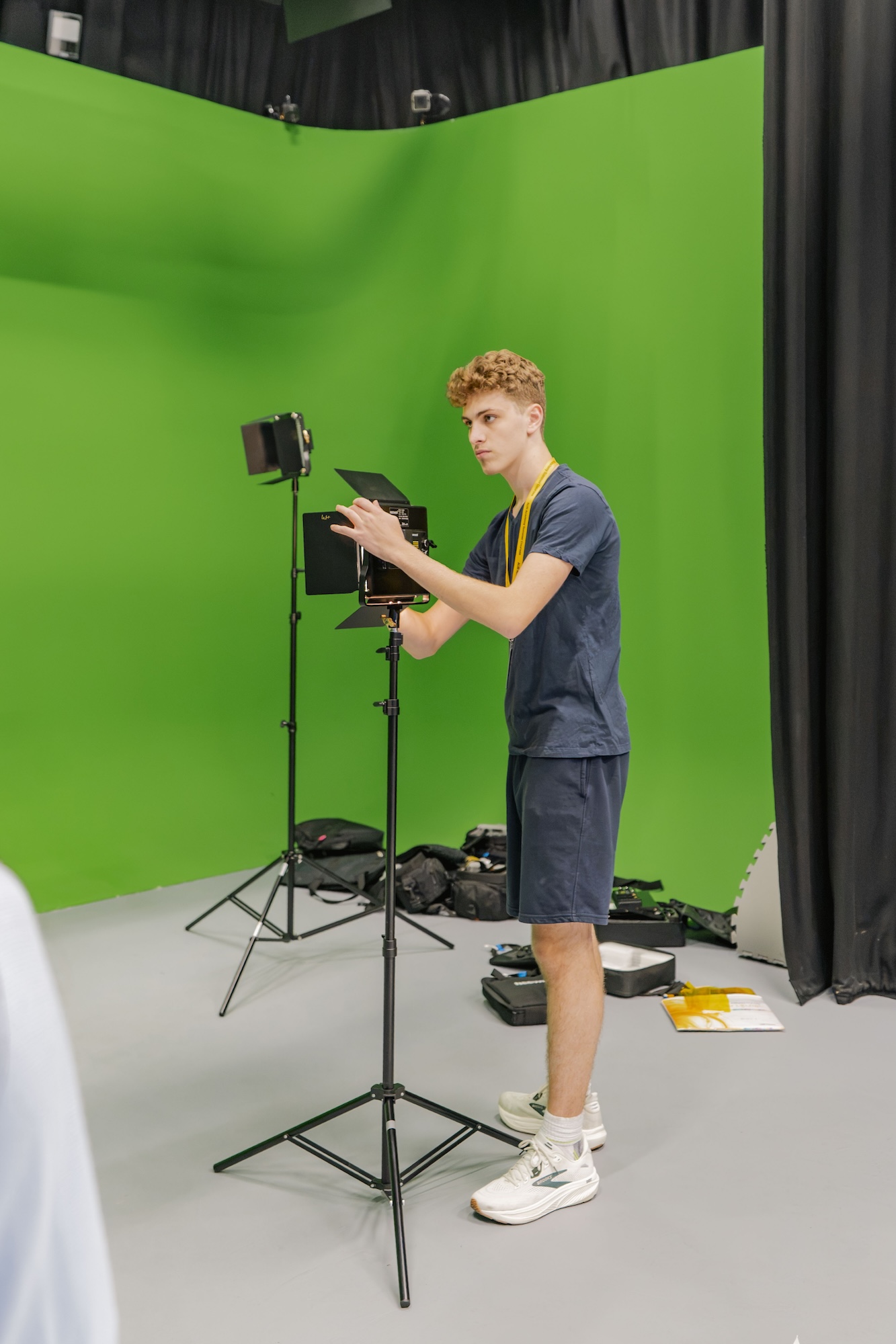 A young man adjusts a tablet on a tripod in a green-screen studio, with a studio light, black curtains and equipment on the floor behind him.