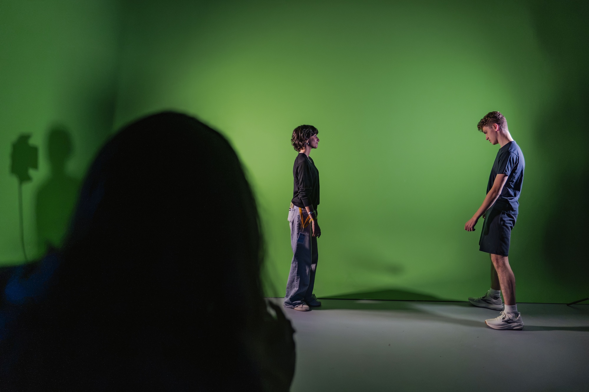 Two people stand facing each other in front of a green screen, with an out-of-focus silhouette in the foreground.