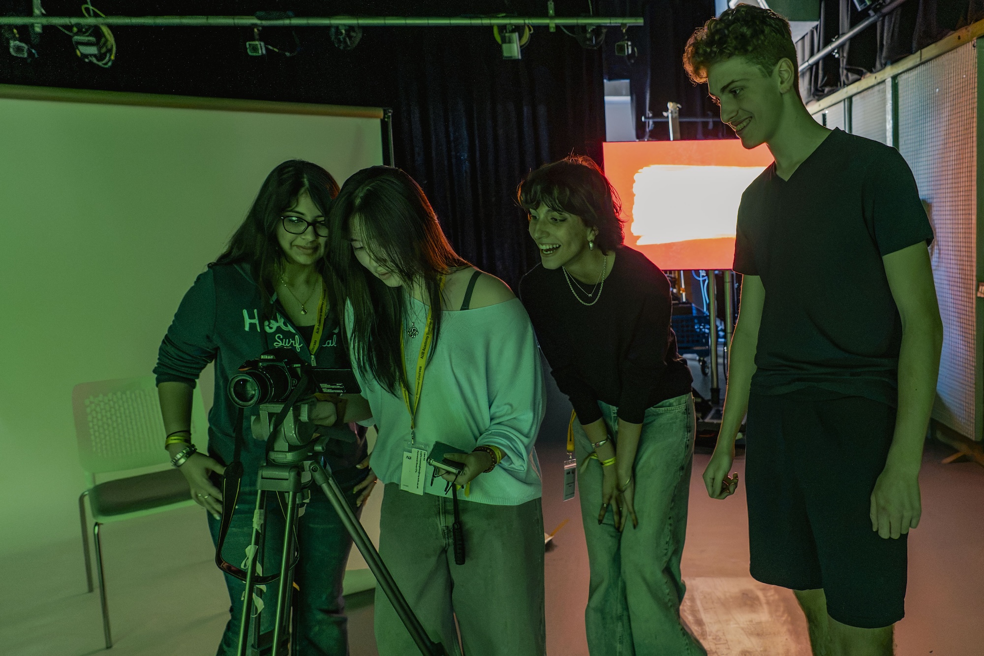 Four young people gathered around a camera on a tripod in a studio, with green and orange lights in the background.