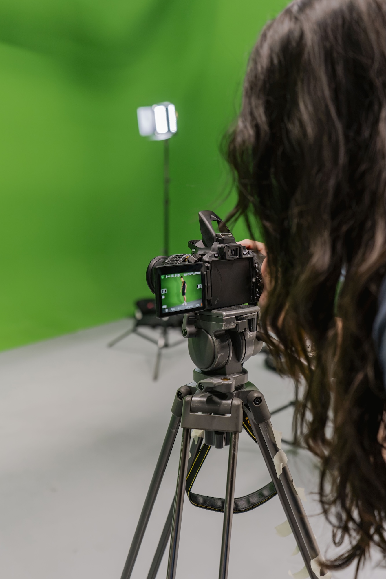 A woman films a camera on a tripod in front of a green screen, with a studio light on a stand in the background.