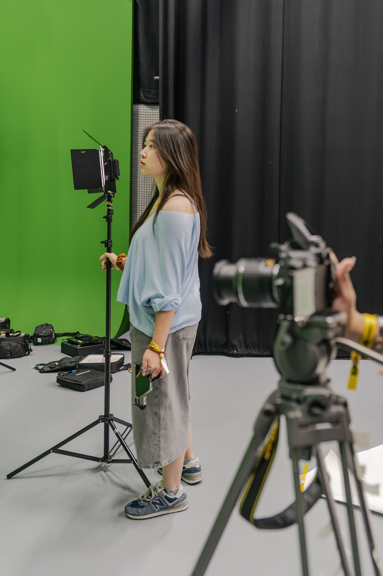 A woman stands beside a studio light with a green screen and black curtains behind her, while a camera on a tripod is visible in the foreground.