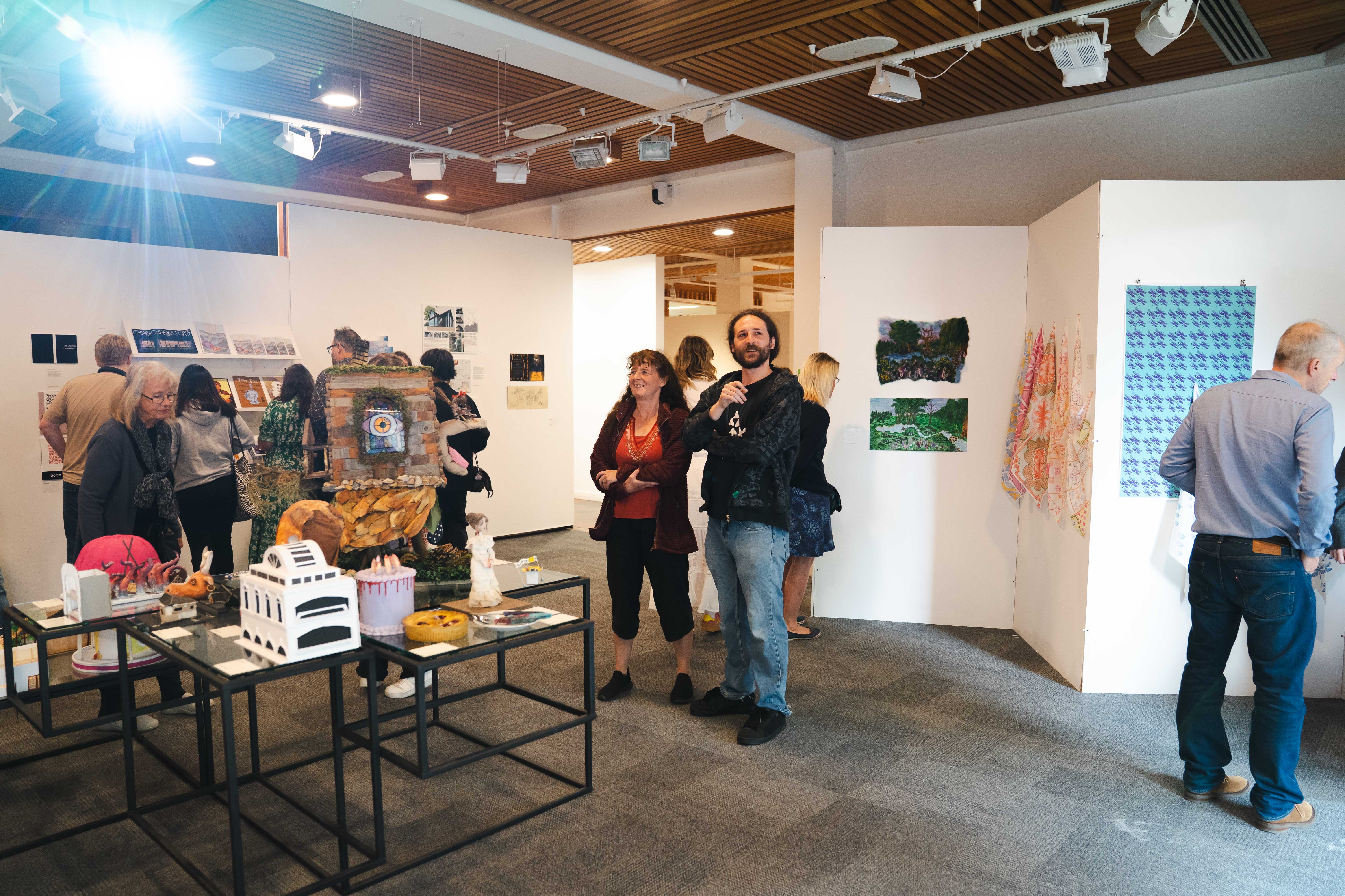 Art gallery interior with visitors viewing wall-mounted works and table displays. White walls, exposed wooden ceiling, concrete floor.