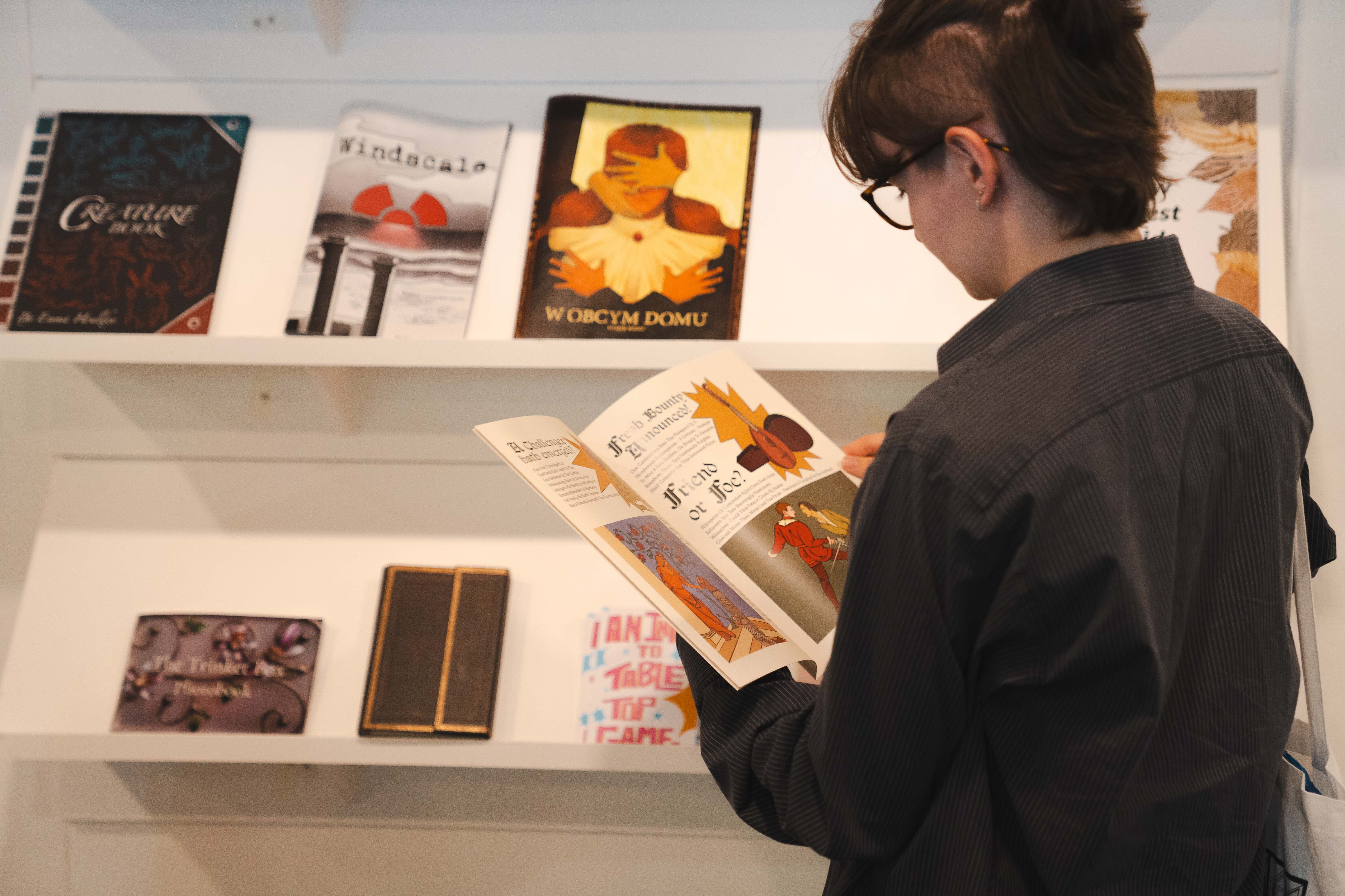 Person with glasses and dark hair reading an open book whilst standing beside white shelves displaying various books and publications.