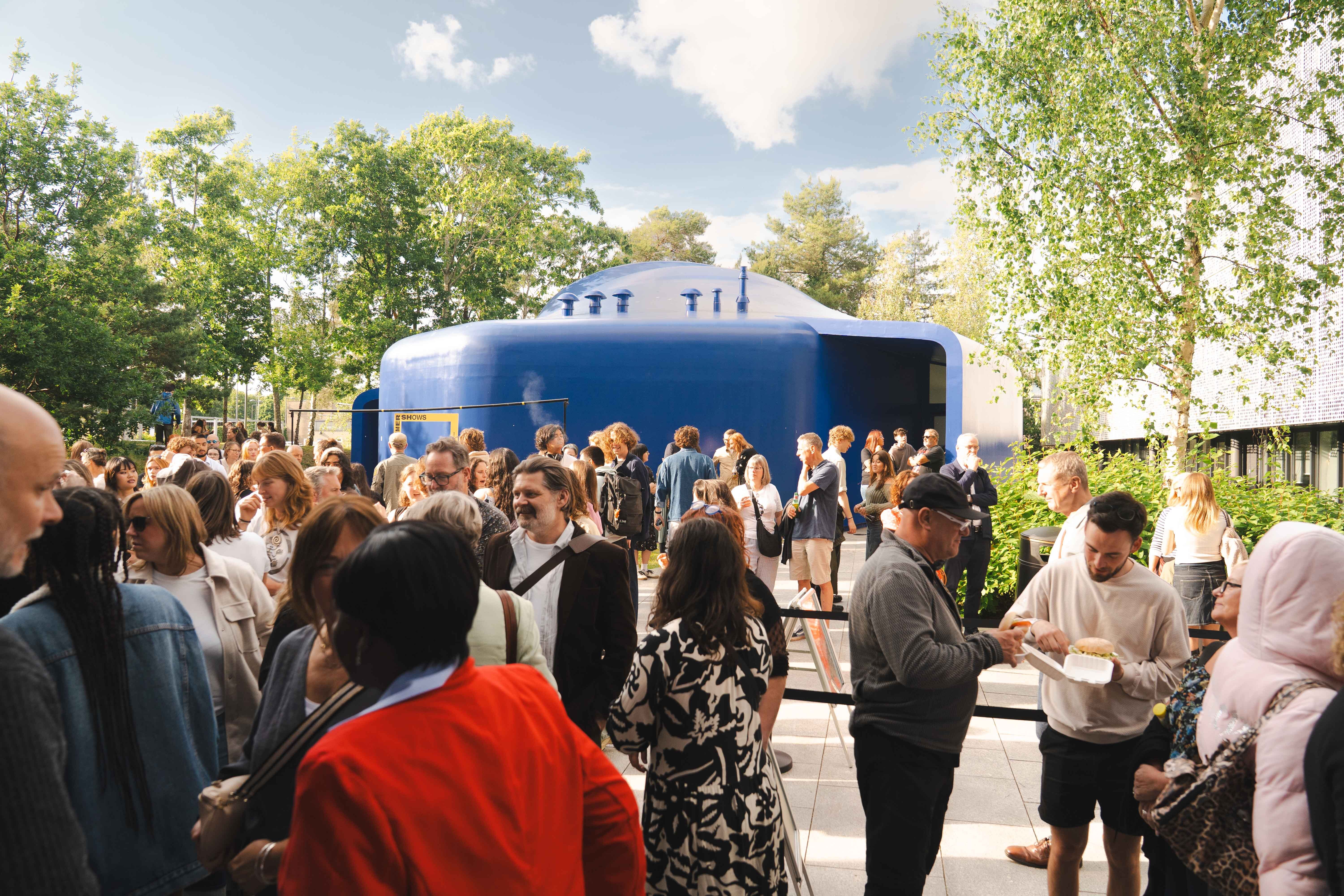 Crowd of people gathered outside blue cylindrical building with white roof under cloudy sky, surrounded by green trees.